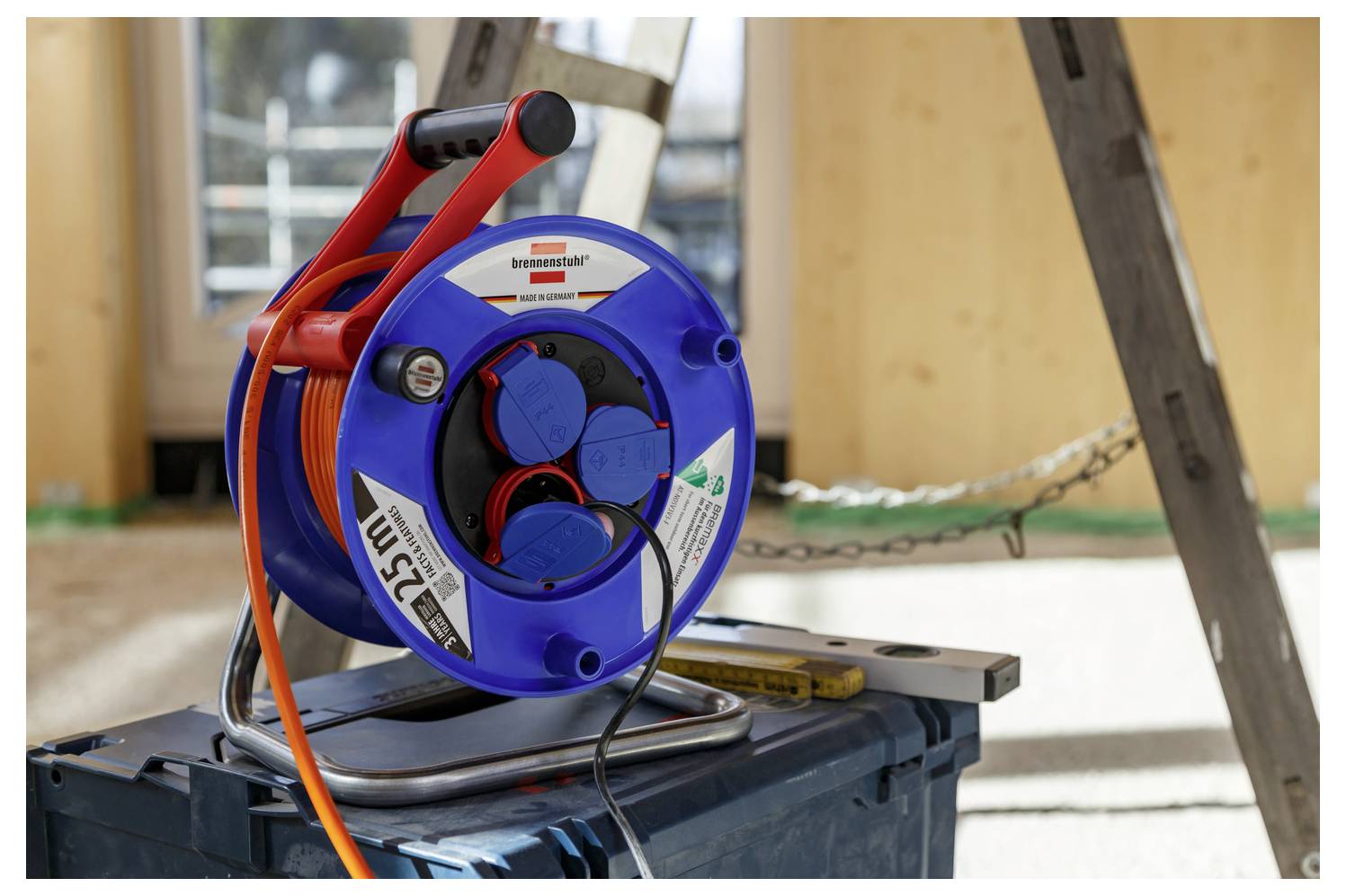 Blauw-en-rood kabelhaspel op een werkbank, met oranje verlengsnoer en stopcontacten, in een werkplaats- of bouwomgeving.