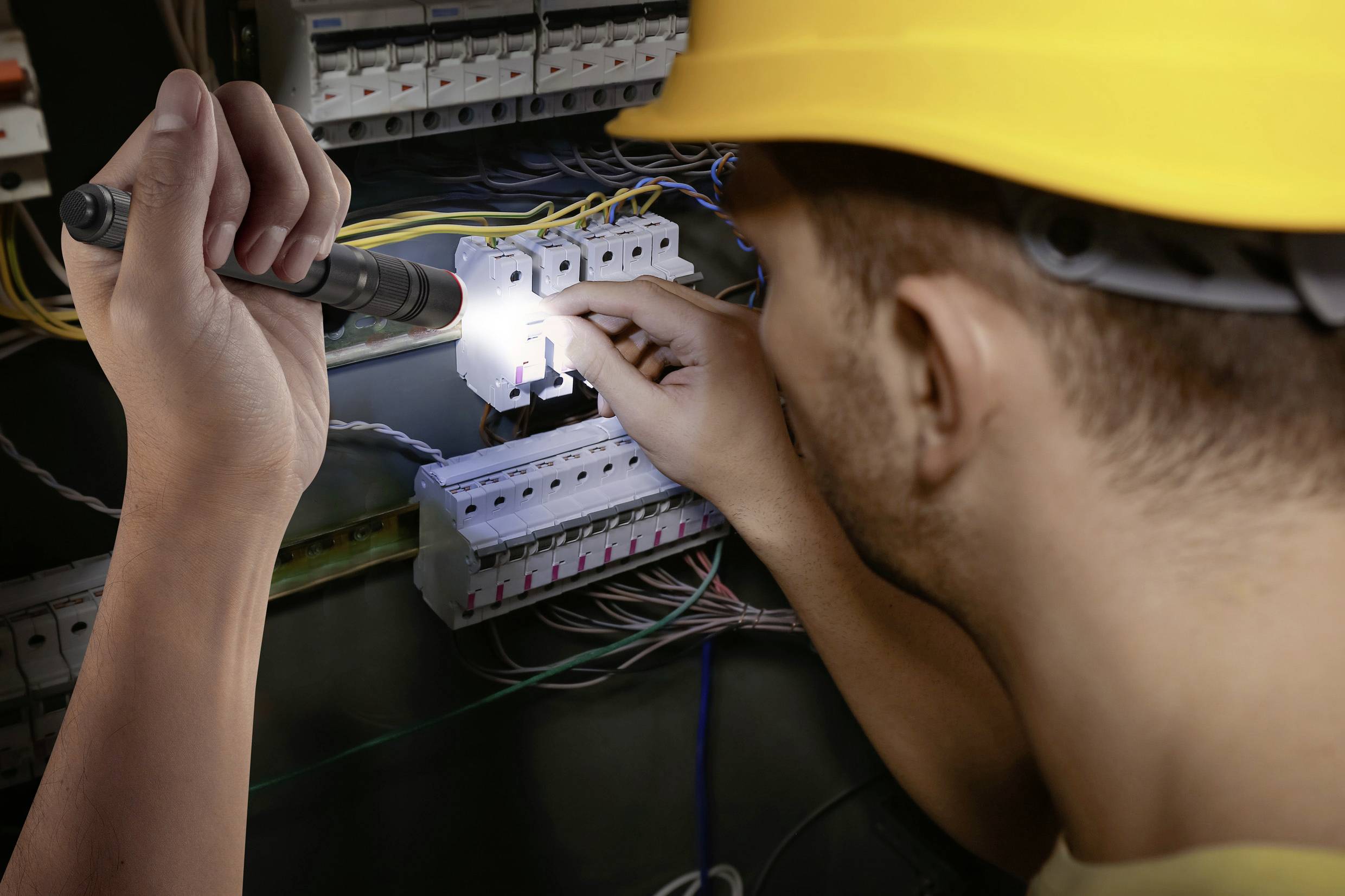 Een elektricien met een gele helm werkt met een zaklamp aan een open zekeringskast met veel draden.