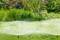Gras in de tuin wordt besproeid door automatische sprinklers. Op de achtergrond staat dicht groen gebladerte.