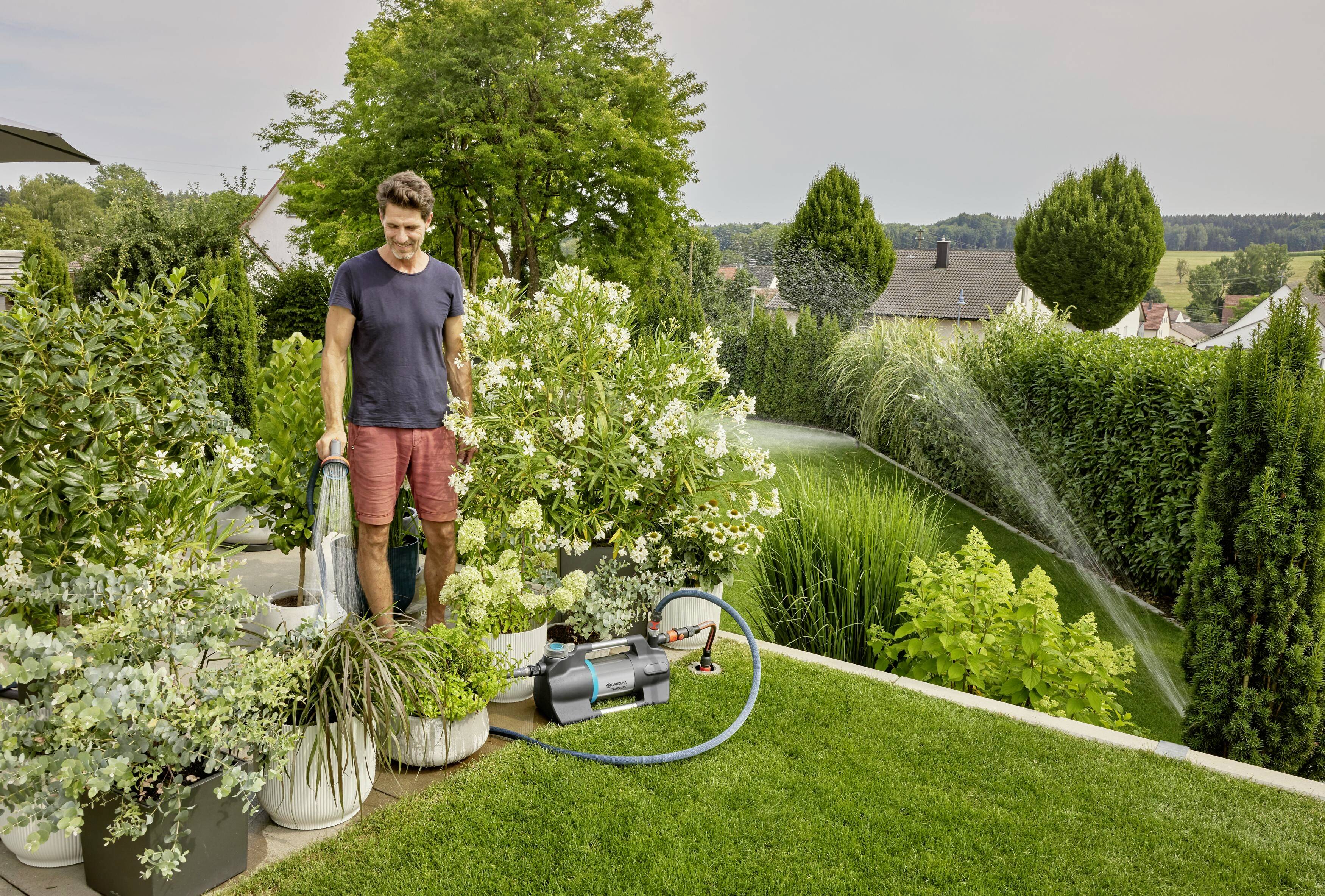Een man besproeit planten in een groene tuin met een tuinslang, omringd door struiken en bomen. Irrigatiesysteem in gebruik.