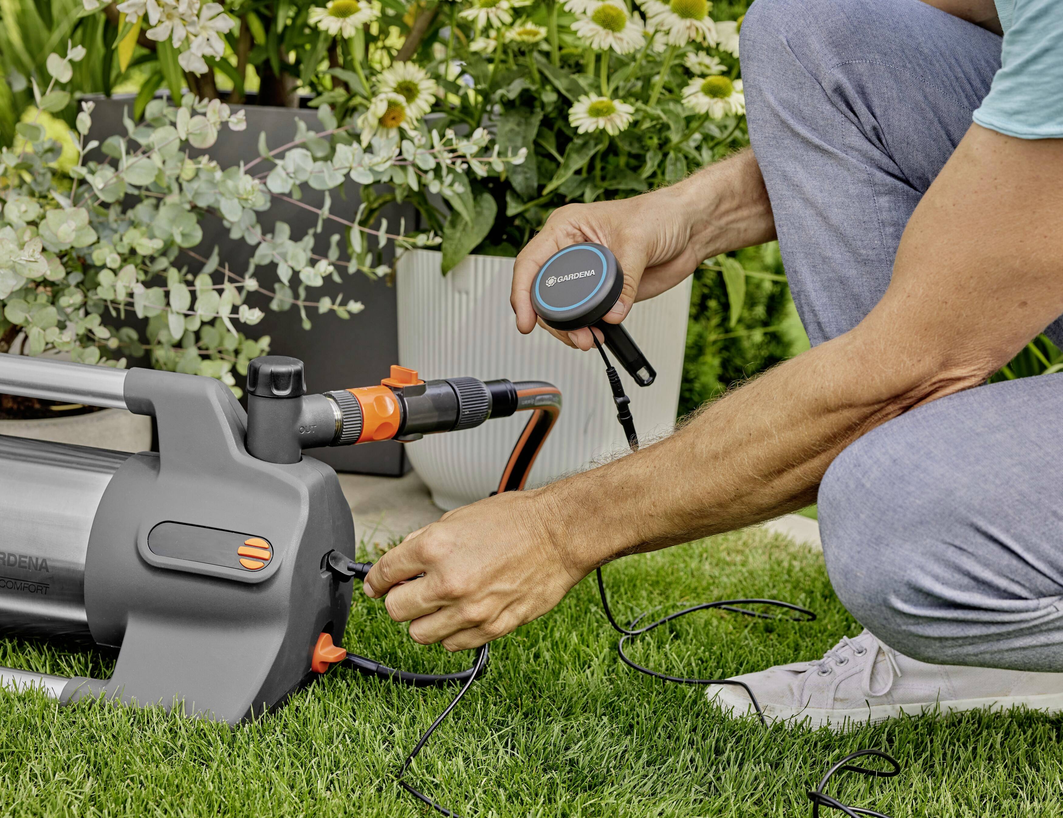 Een persoon installeert een tuinslangpomp op een gazon, omringd door planten. De focus ligt op de handen en de techniek.