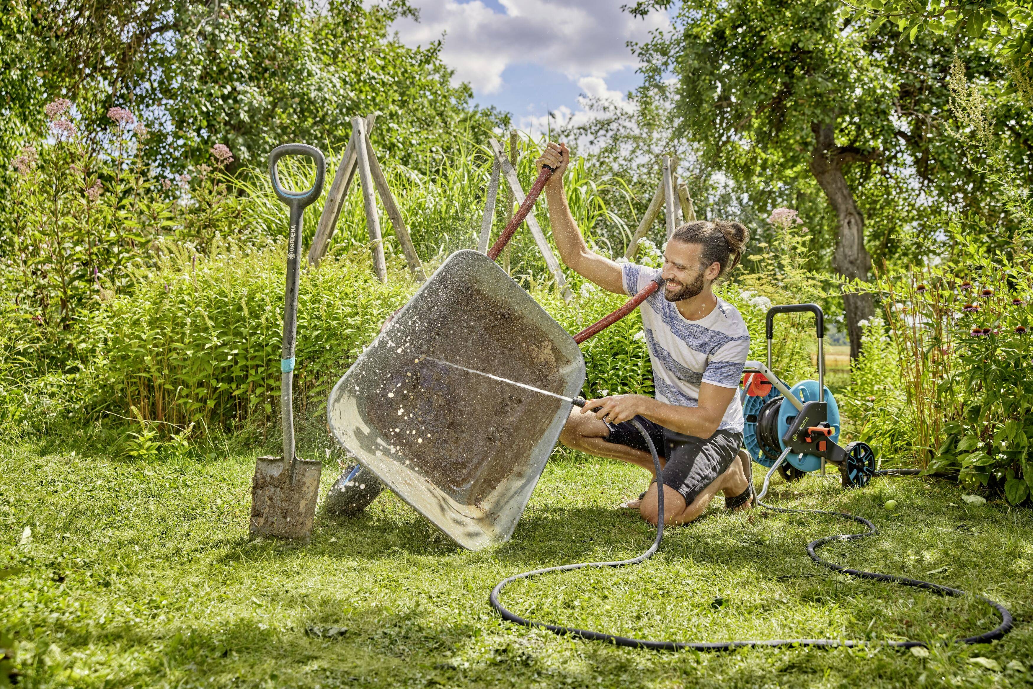 Een man reinigt een kruiwagen met een slang in de zonnige tuin. Naast hem staan een schop en een waterslang.