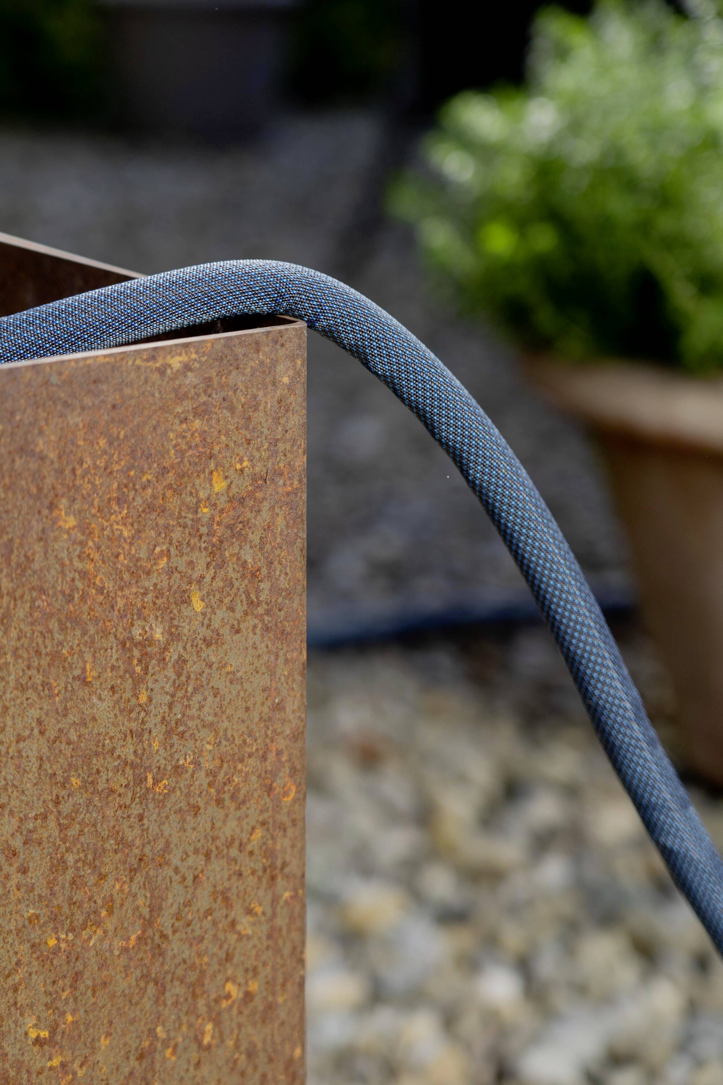 Een grijze tuinslang hangt over een roestige metalen plaat buiten. Op de achtergrond zijn onscherpe planten zichtbaar.