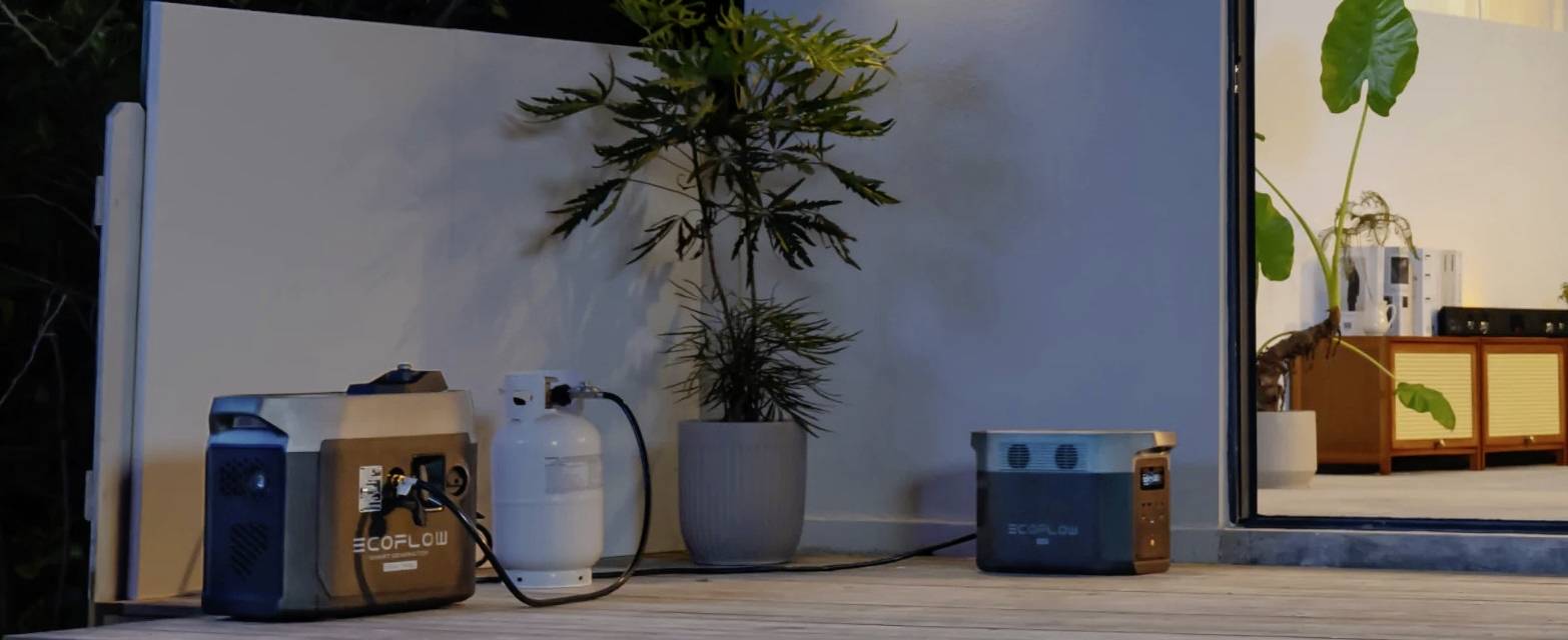 Een draagbare accu en een gasfles staan op een terras naast een plant, verbonden door een kabel. Op de achtergrond is binnenverlichting zichtbaar.