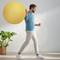 Een man in een blauwe trui houdt een grote gele fitnessbal vast. Naast hem staat een plant in een pot voor een witte bakstenen muur.