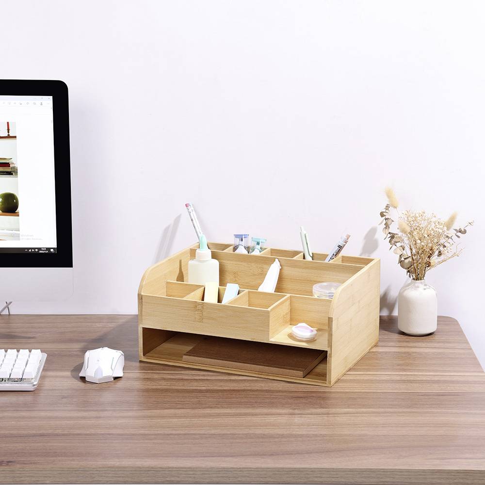 Een bureau met een computerscherm, een toetsenbord en een houten organizer-box met schrijfbenodigdheden. Ernaast een vaas met takken.