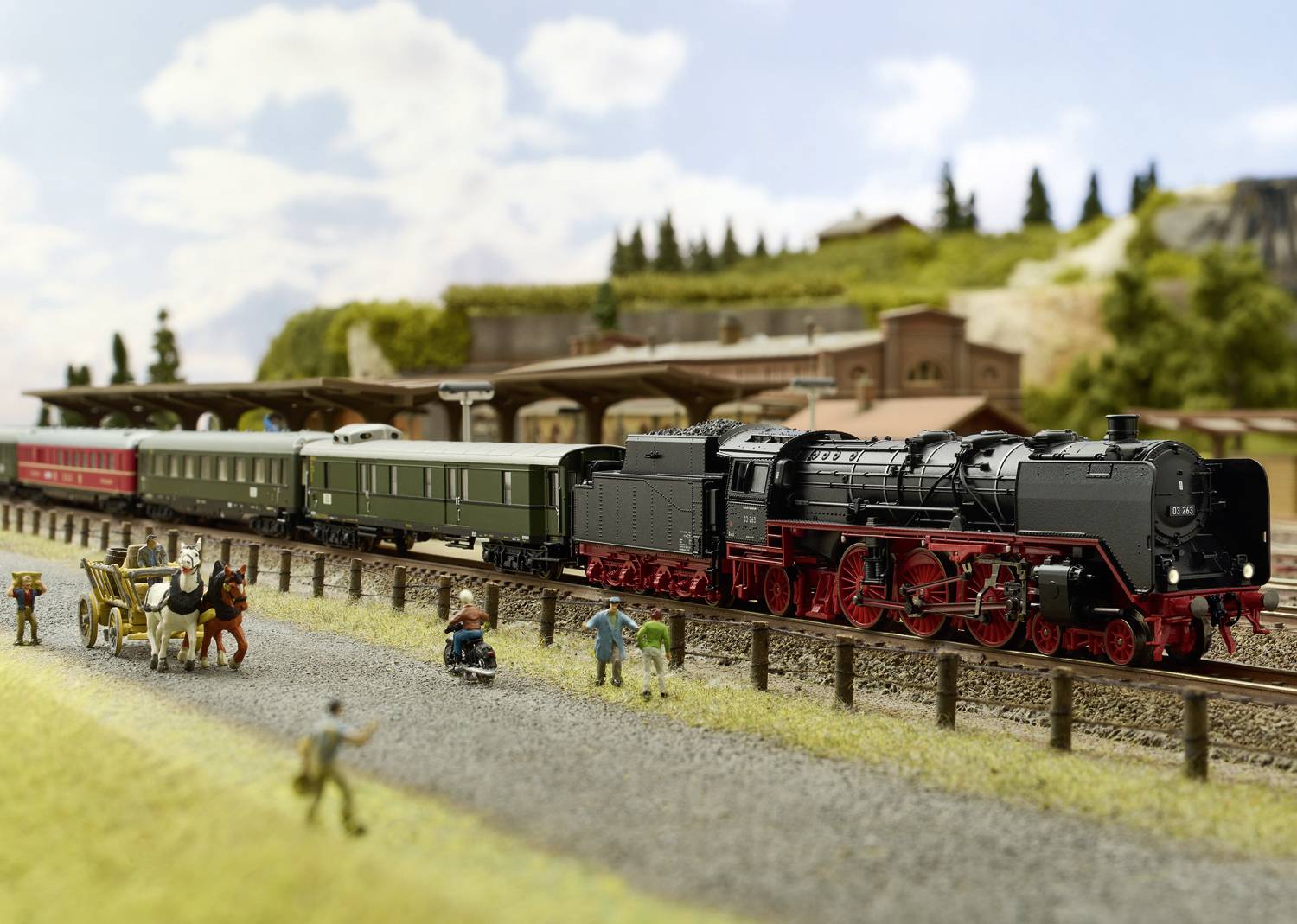 Modelspoorbaan met een zwarte stoomlocomotief en personen op het perron. Een koets rijdt voorbij, gebouwen op de achtergrond.