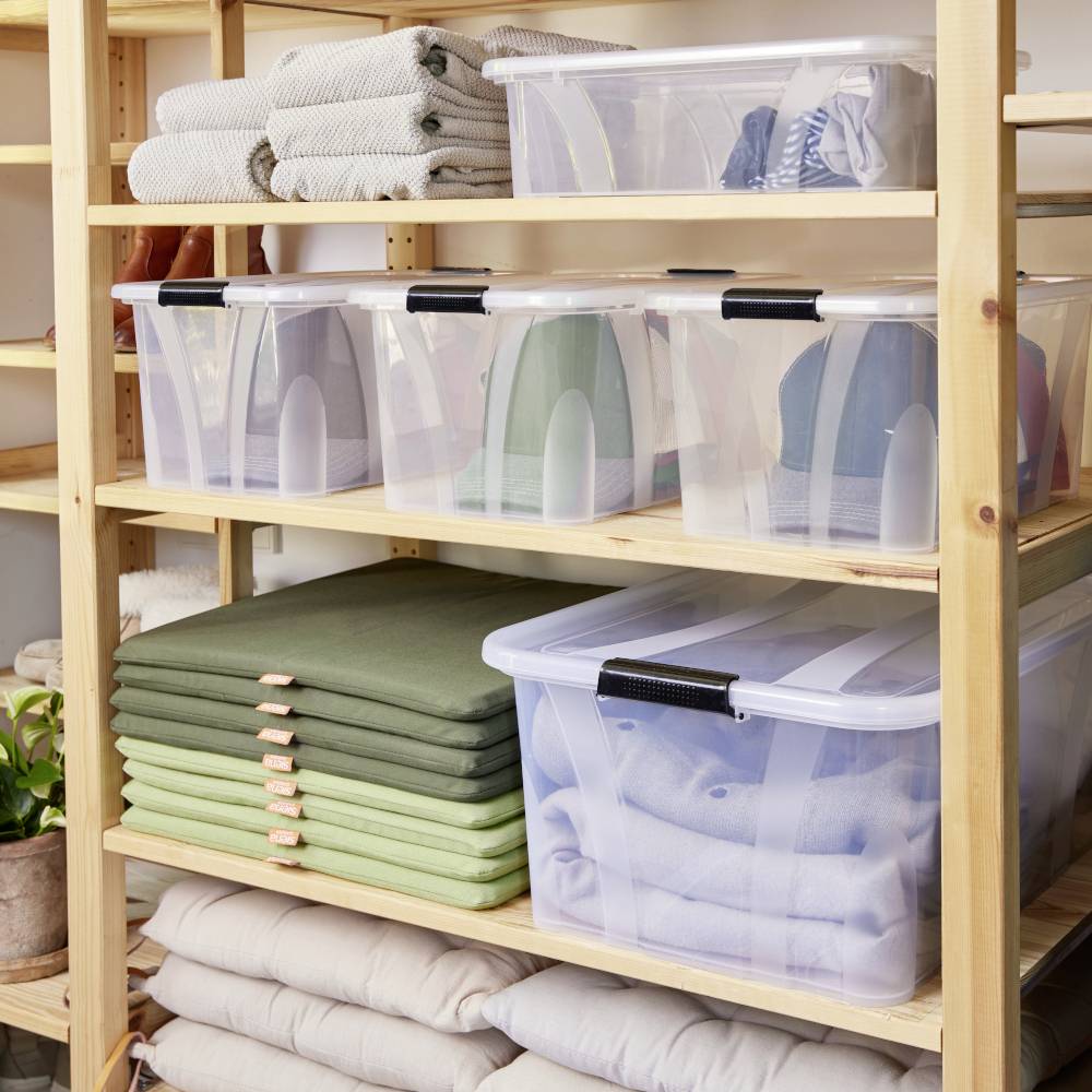 Een houten rek met plastic dozen en gevouwen dekens. Bovenaan gevouwen handdoeken, onderaan opslagdozen met kleding. Een plant aan de linkerkant.