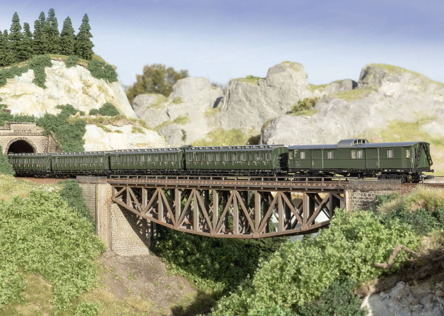 Een groene modeltrein rijdt over een spoorbrug in een landschap met bergen en bomen op de achtergrond.