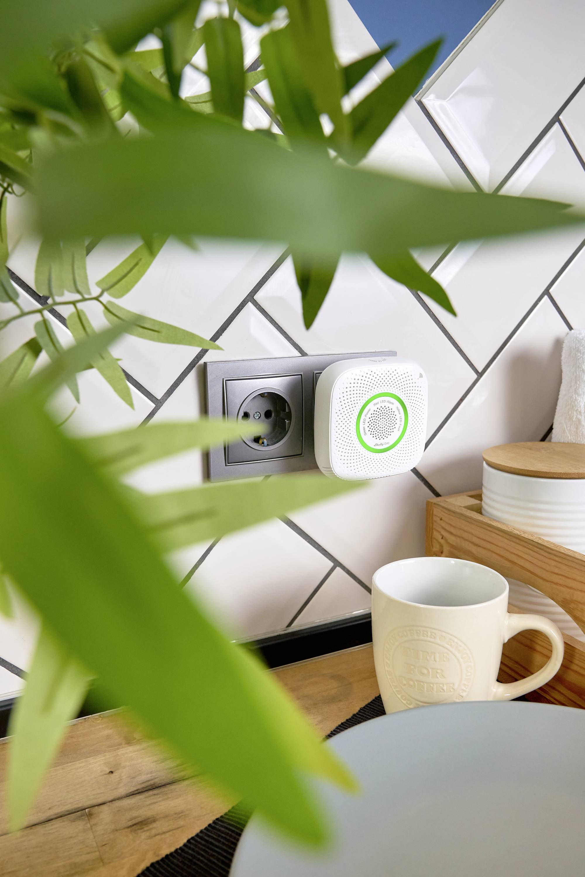 Een kleine witte luchtkwaliteitsmonitor met een groene LED-ring is in een stopcontact in een keuken gestoken, naast een houten blok.