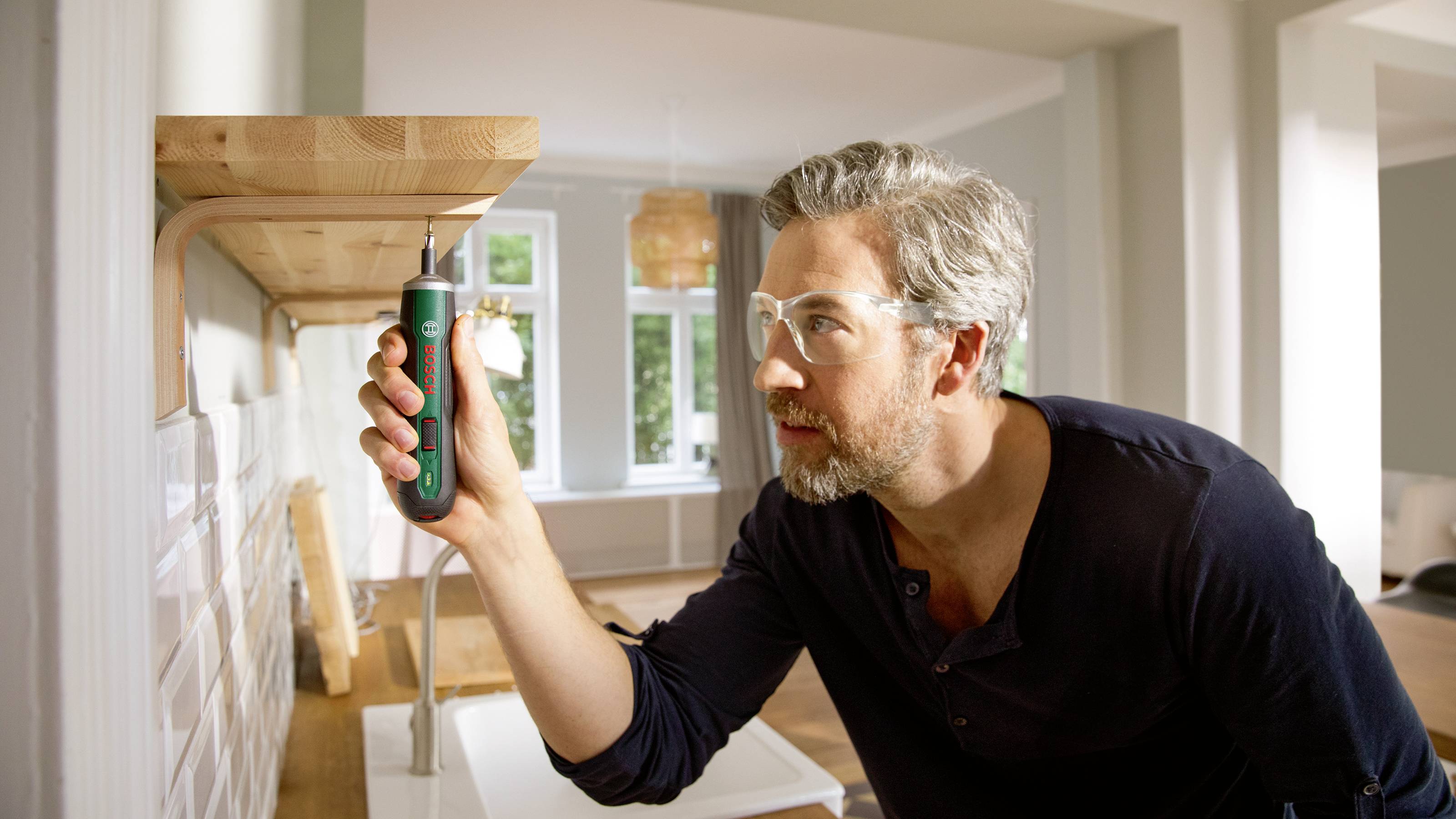 Een man bevestigt een plank aan de muur met een schroevendraaier in een lichte ruimte met grote ramen.
