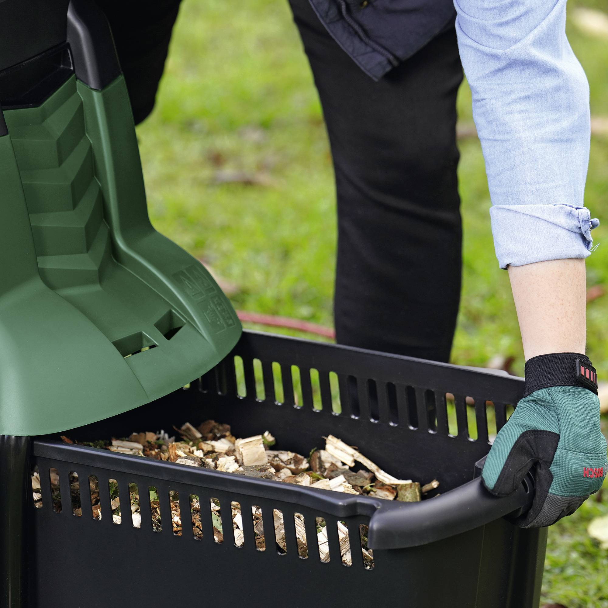 Een persoon met blauwe handschoenen leegt verkleind hout in een zwarte container buiten.