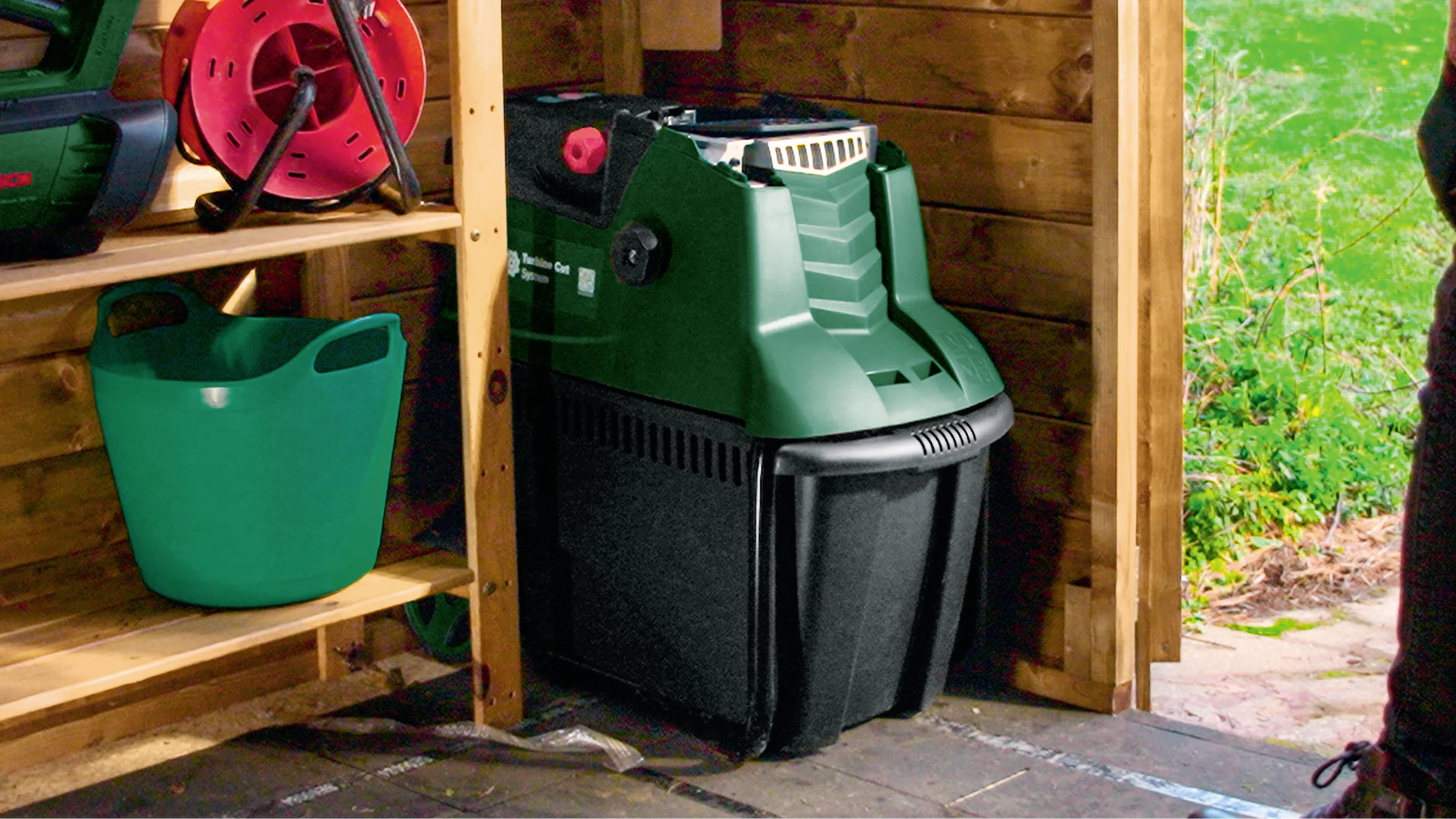 Groene tuinhakselaar staat in een houten schuur. Ernaast is een groene emmer en een tuinslang aan de muur bevestigd. Op de achtergrond is gazon zichtbaar.