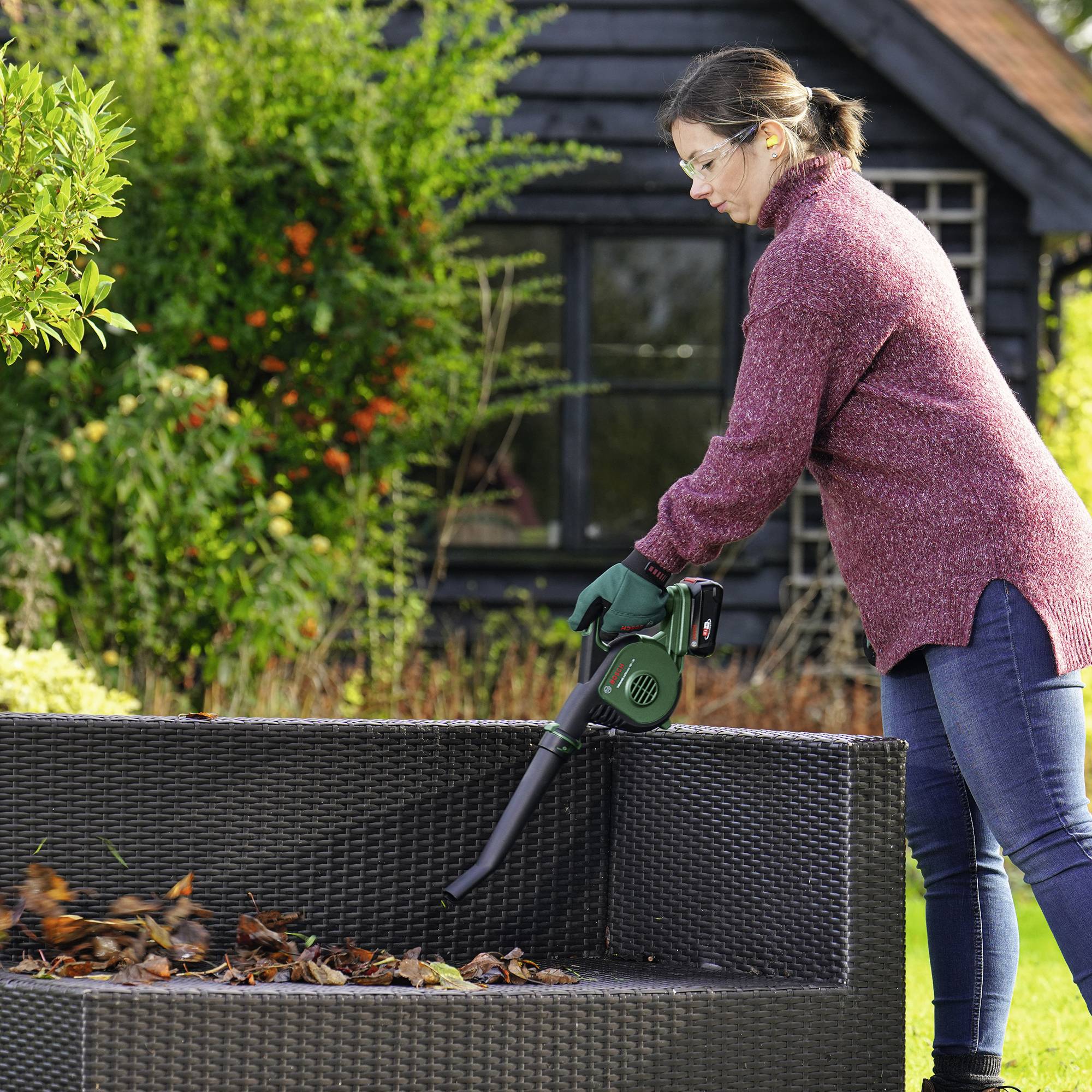 Een vrouw gebruikt een bladblazer om herfstbladeren van een terras in een tuin te verwijderen. Op de achtergrond staat een huis.