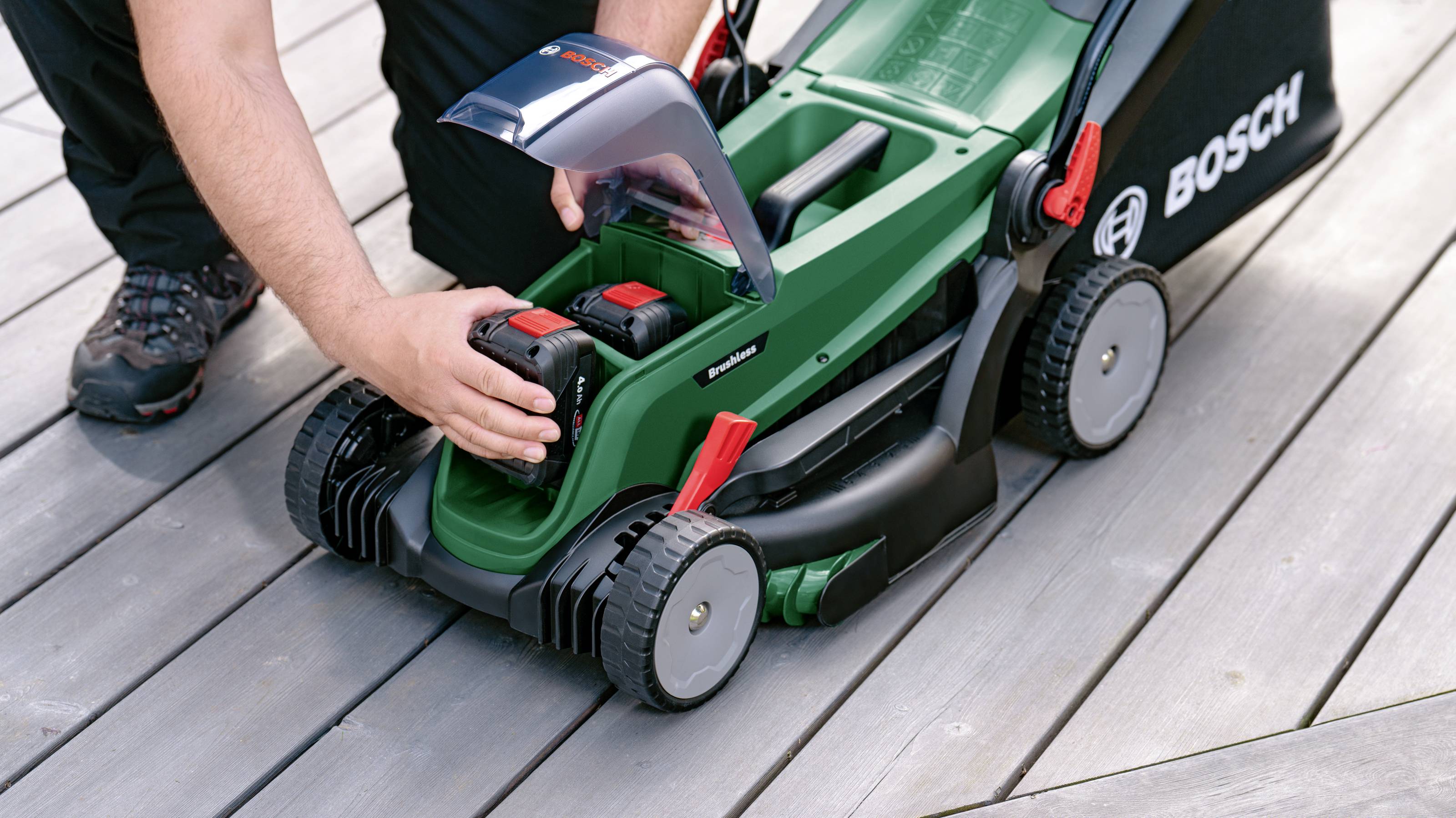 Een persoon houdt de accu van een Bosch-grasmaaier vast terwijl hij deze op een houten terras verwisselt.