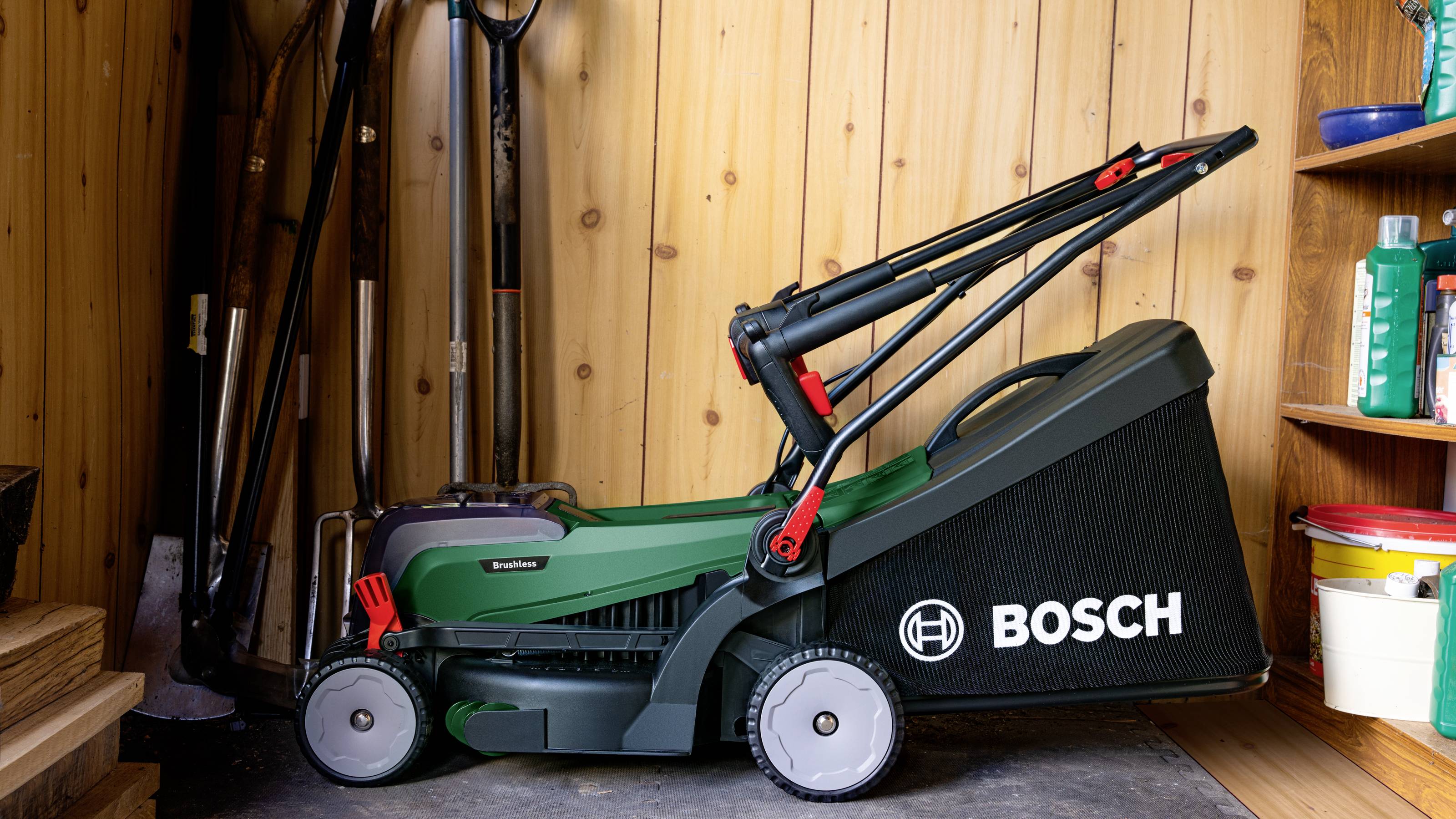 Een Bosch-grasmaaier staat in een houten schuur, omringd door tuingereedschappen en voorraadrekken.