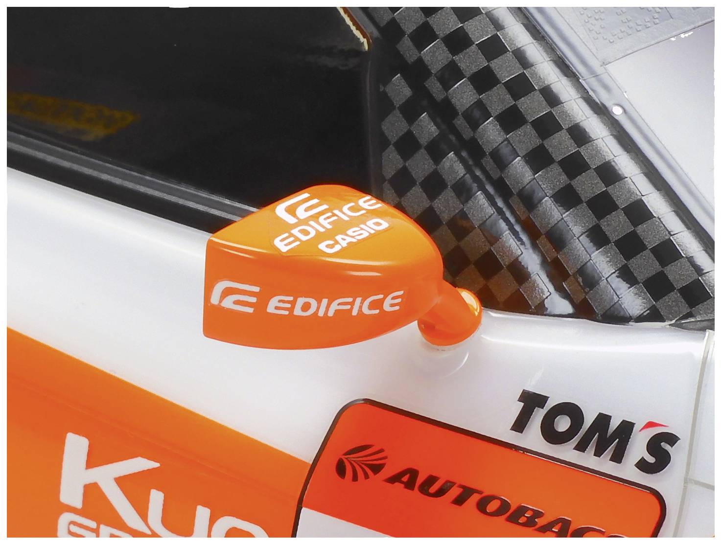 Een oranje zijspiegel van een racewagen met de logo's 'EDIFICE' en 'CASIO', naast 'TOM'S' op een grijs geruiteerd patroon.