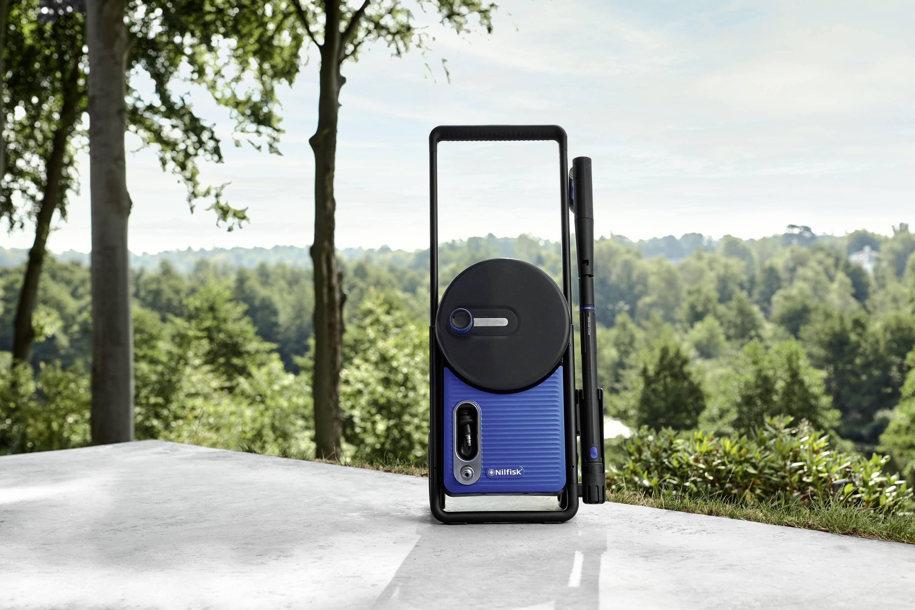 Een draagbaar, blauw apparaat met een zwarte ronde antenne staat op beton. Op de achtergrond is een met bomen bedekte heuvel te zien.