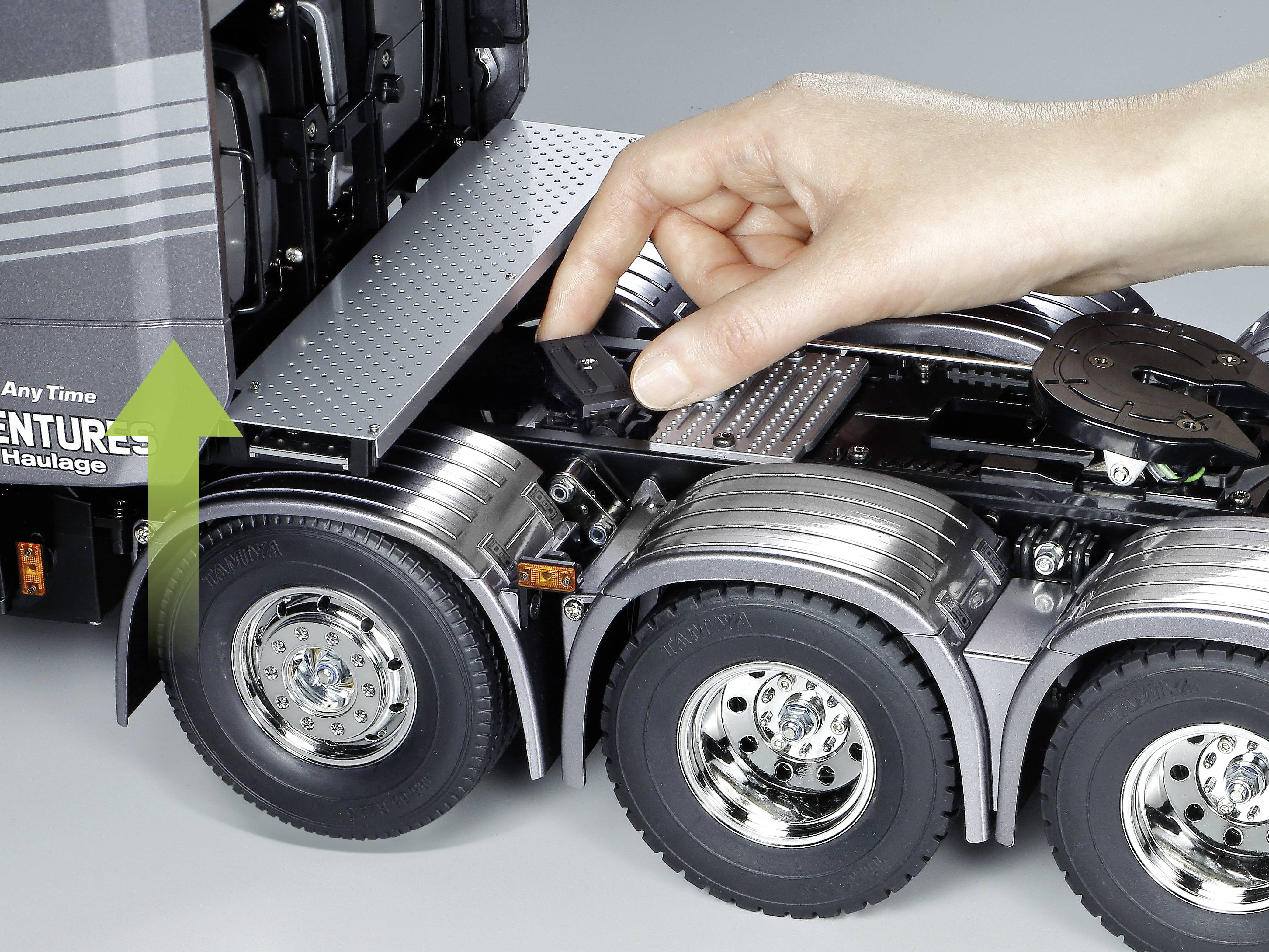 Een hand bedient de knop van een model-vrachtwagen. Een groene pijl wijst naar de achterkant van het voertuig, mogelijk om deze te openen.