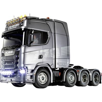 Grijze Scania-vrachtwagen zonder aanhanger, vier assen, rijdt op een natte weg. Moderne cabine, lichten branden. Bestuurder zichtbaar binnenin.