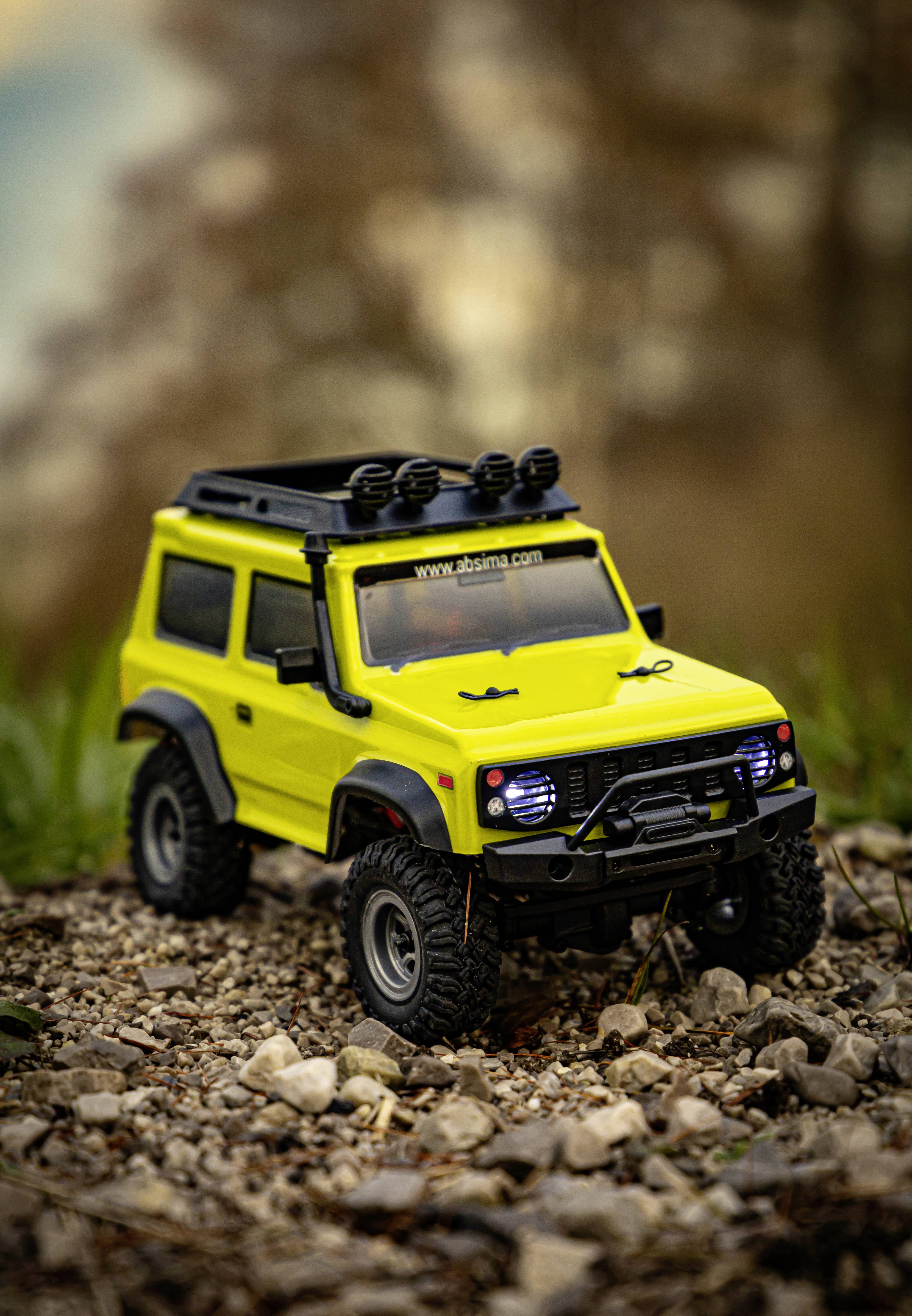 Geel modelauto van een terreinwagen met zwarte details staat op een stenige ondergrond buiten, achtergrond wazig.