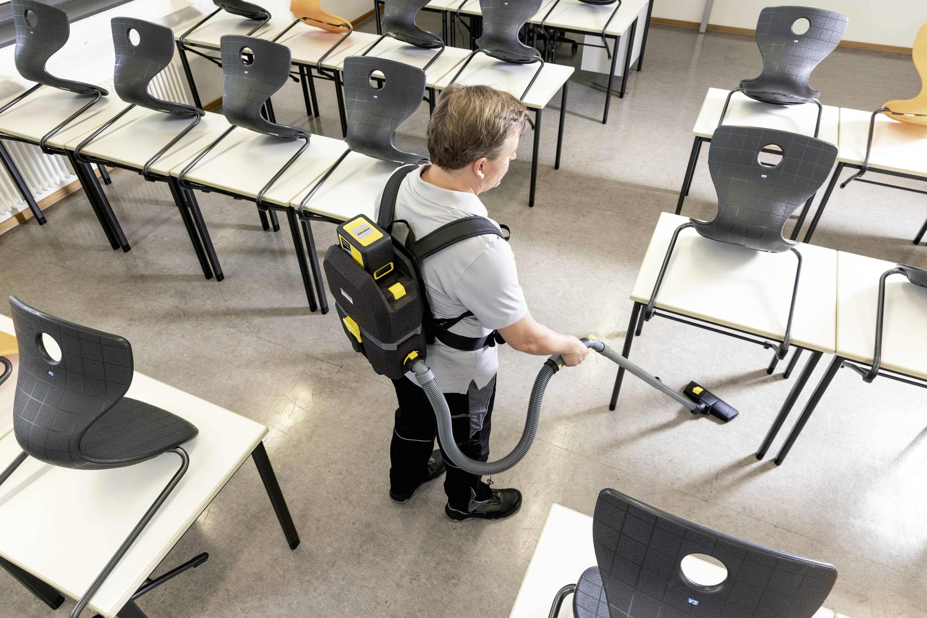 Man met rugzakstofzuiger reinigt een lege klaslokaal. Stoelen zijn omhoog gezet, de vloer wordt gestofzuigd.