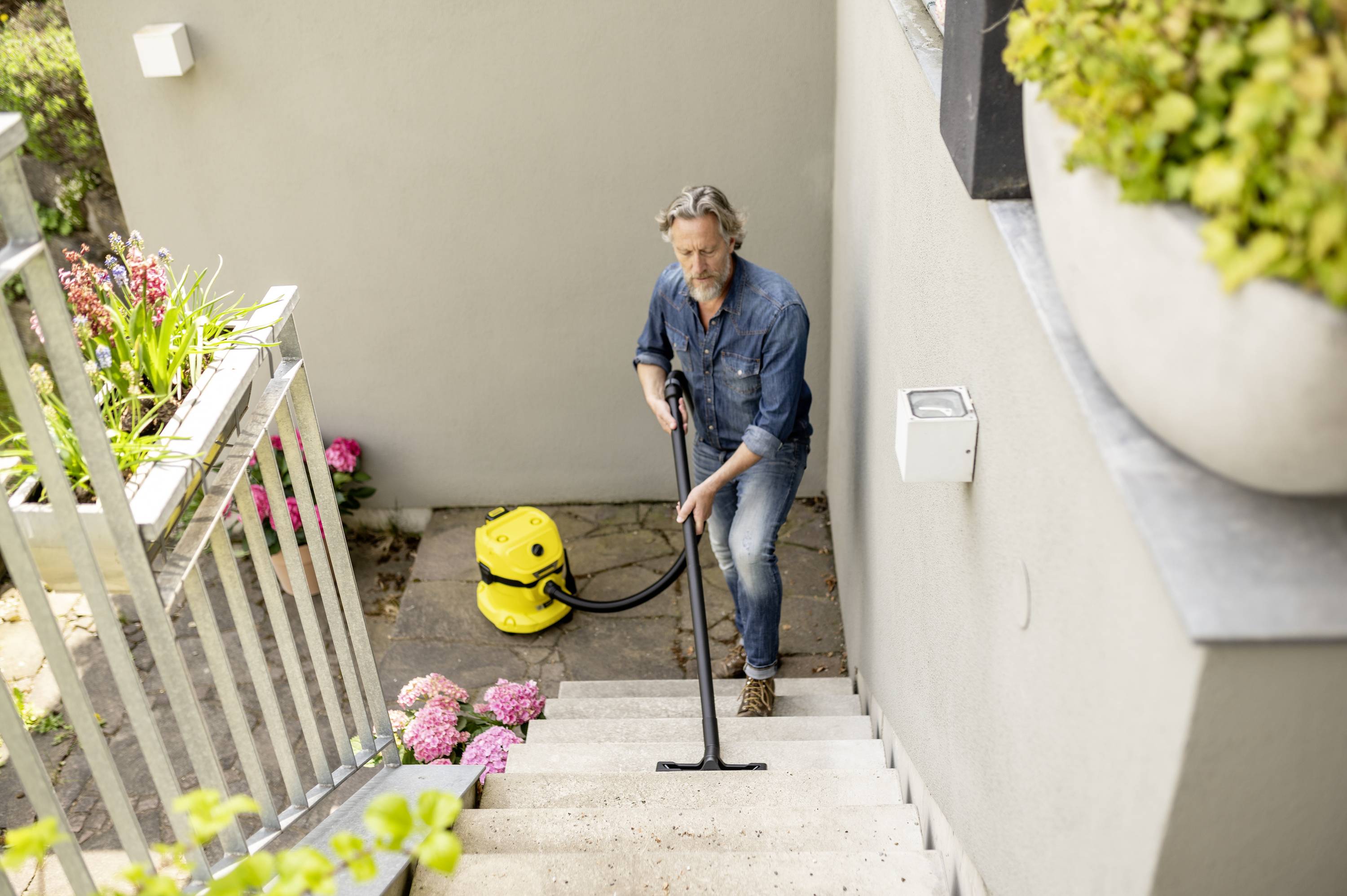 Een man reinigt een trap buiten met een gele stofzuiger. Naast hem bloeien roze hortensia's en groene planten.