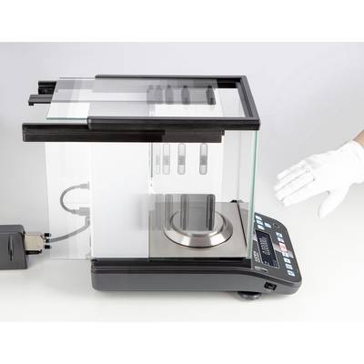 Een laboratoriumweegschaal met een glazen windscherm toont een hand in een witte handschoen die voorzichtig dicht bij de weegschaal wordt gebracht.