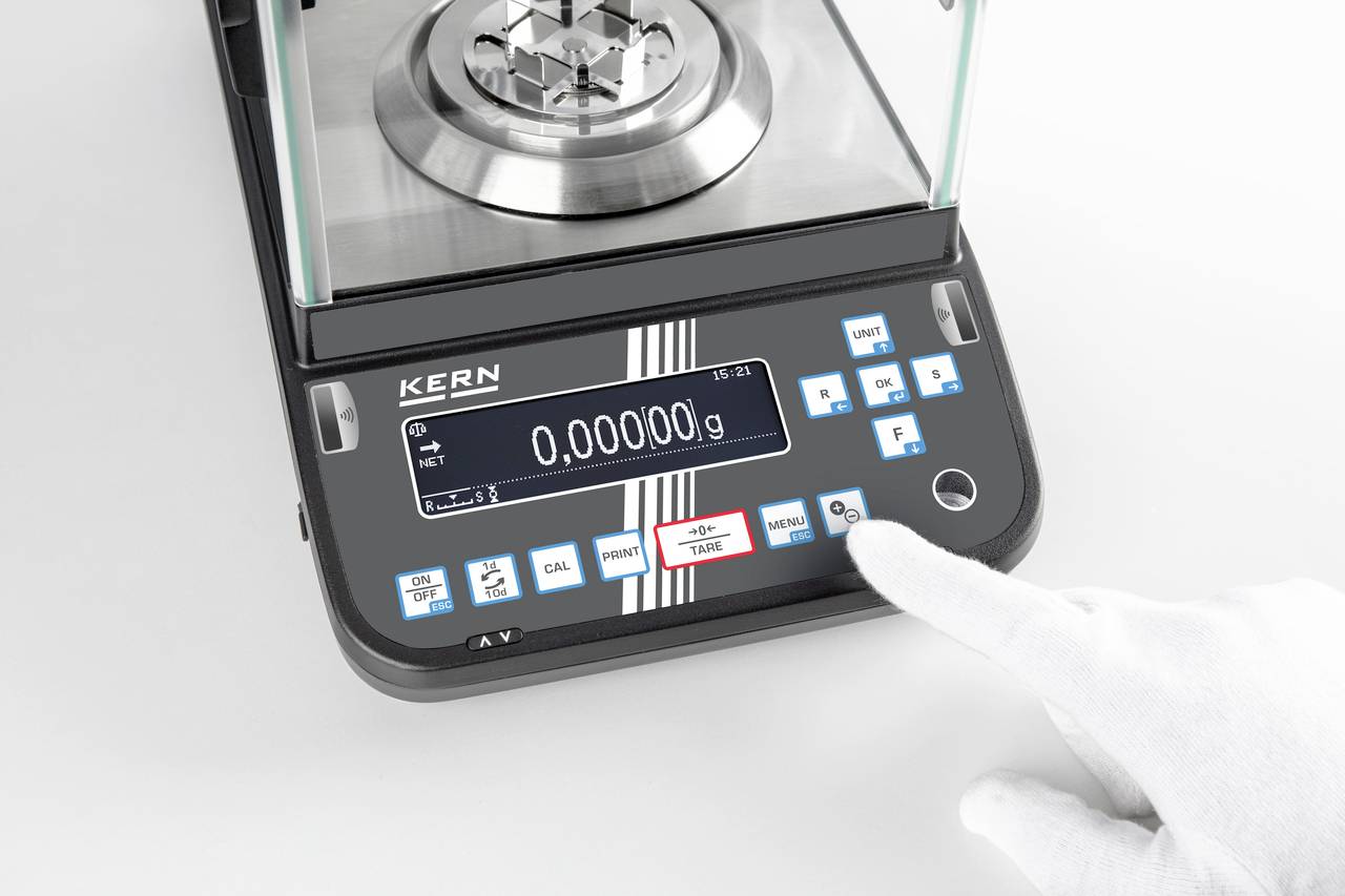 Een elektronische weegschaal met een digitaal display toont '0,00000 g'. Een handschoen drukt een toets om het menu te selecteren.