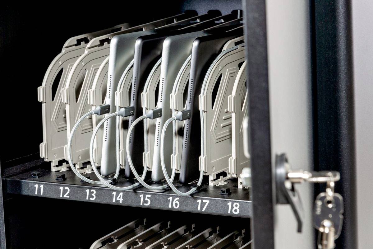 Een open metalen kast toont veilig opgeslagen laptops op genummerde vakken, met oplaadkabels aangesloten.
