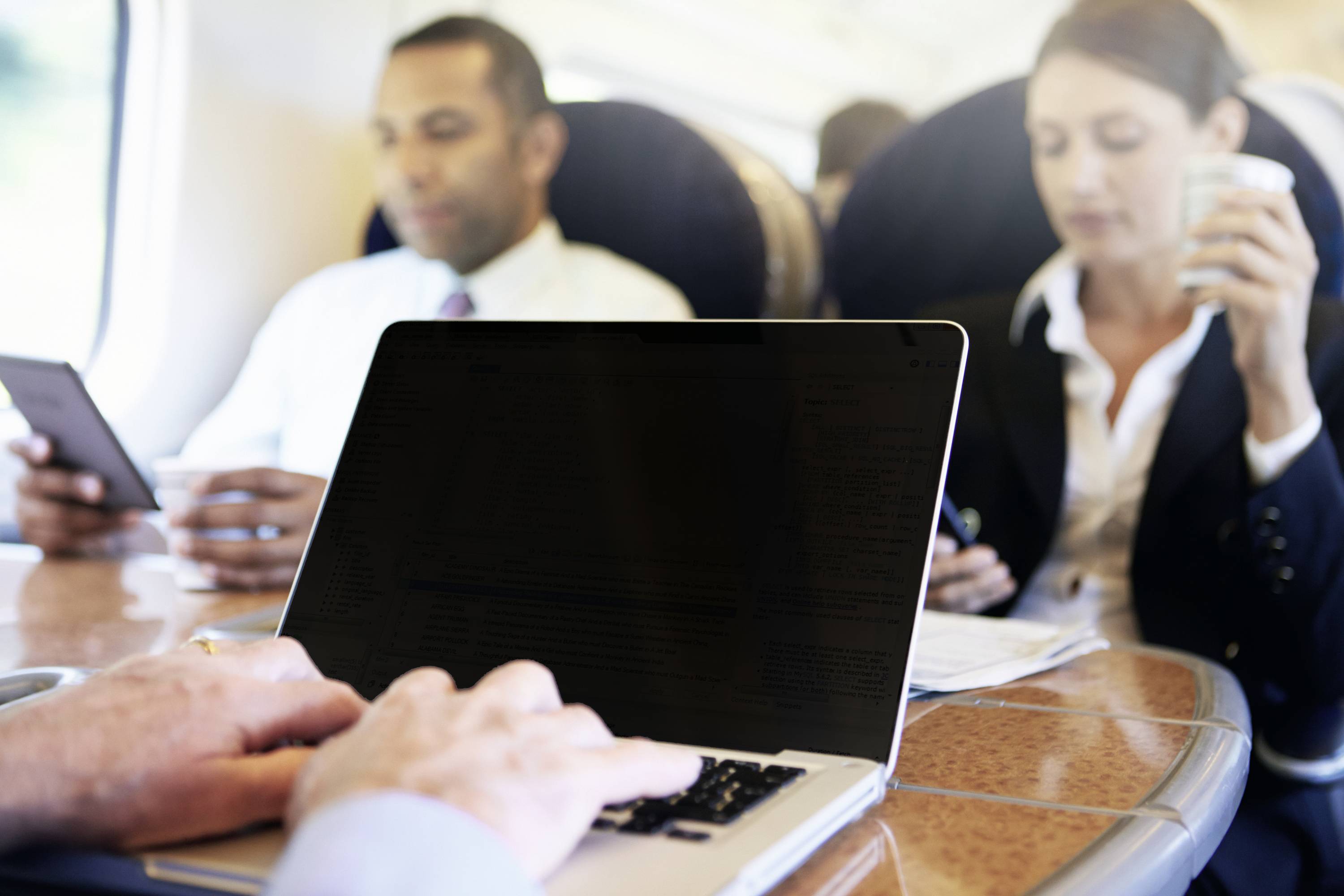 Een persoon werkt op een laptop in een trein, terwijl twee andere personen op de achtergrond een tablet en een koffiekopje vasthouden.
