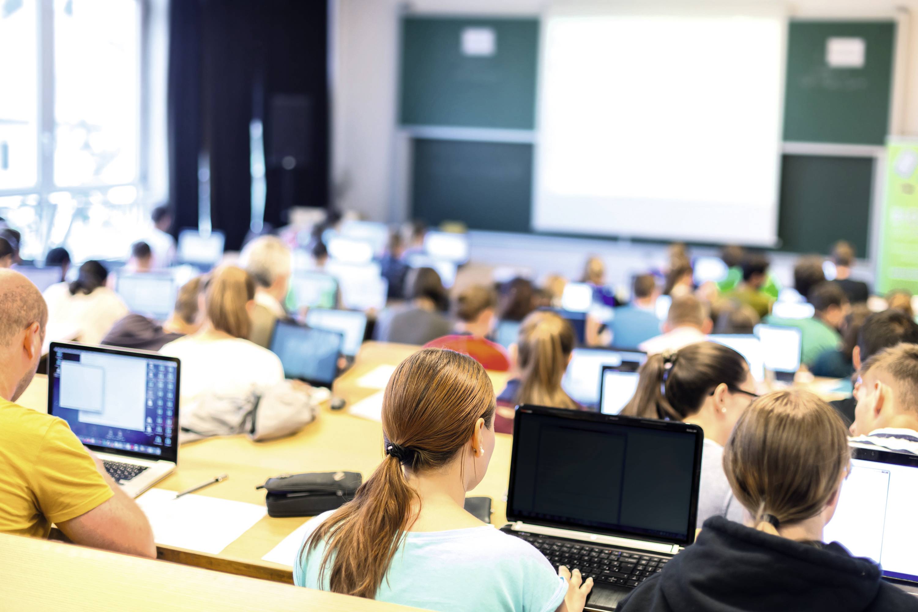 Studenten zitten aan tafels in een klaslokaal en werken op laptops. Op de achtergrond zijn schoolborden en een beamer zichtbaar.