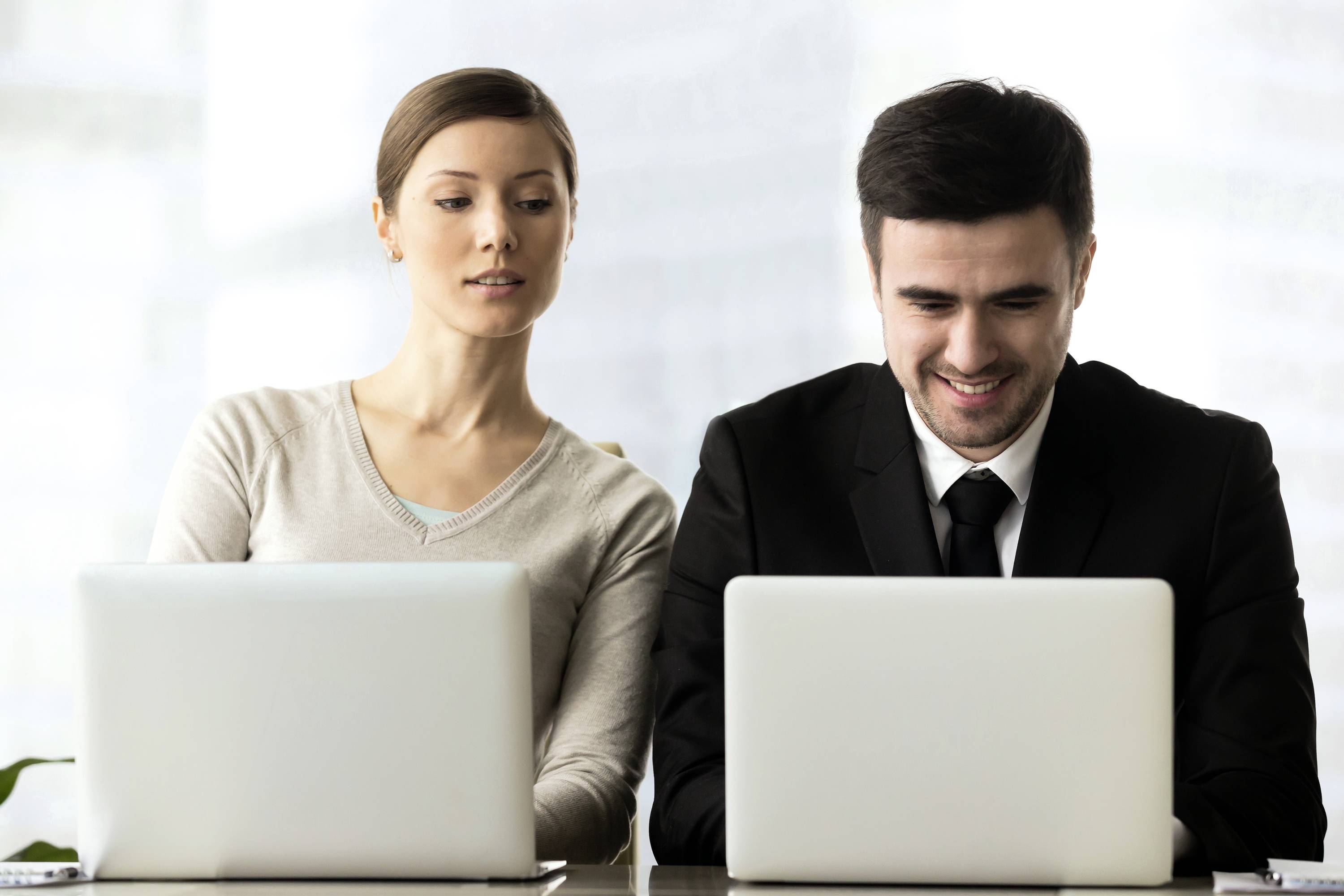 Een vrouw en een man, beiden zittend achter laptops. De man glimlacht naar zijn scherm, terwijl zij geïnteresseerd naar zijn beeldscherm kijkt.
