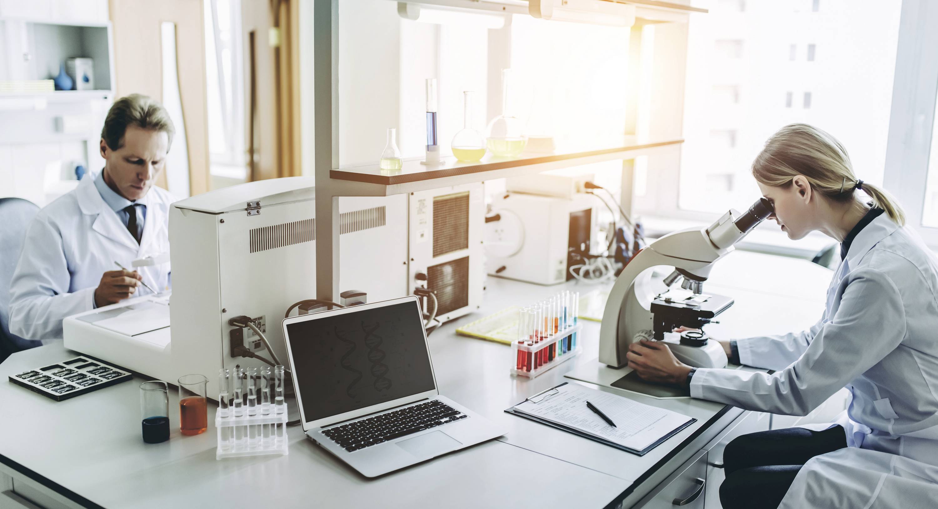 Twee wetenschappers werken in een laboratorium. Een persoon gebruikt een microscoop, terwijl de ander resultaten noteert. Verschillende laboratoriumapparatuur is zichtbaar.