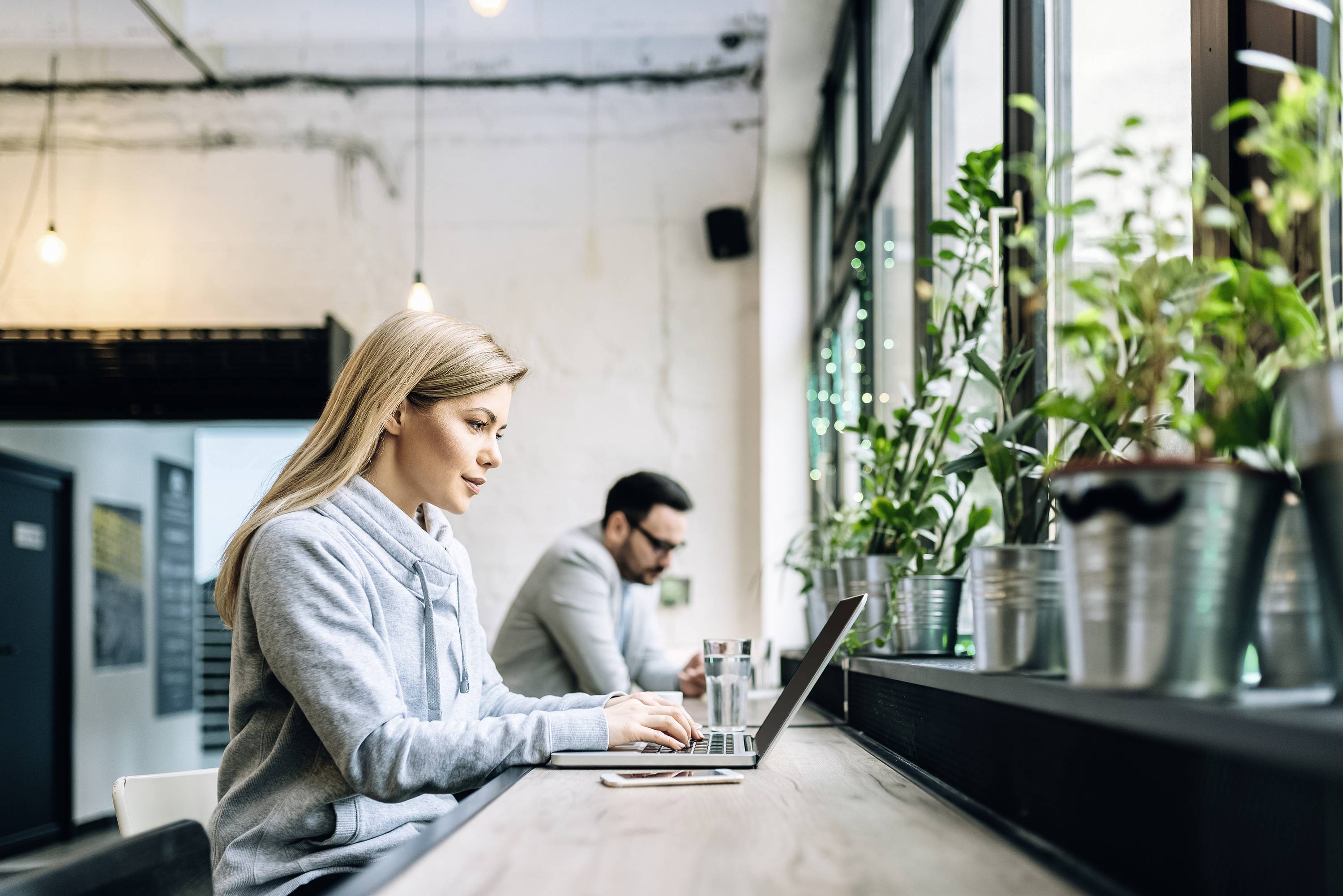 Een vrouw en een man werken op laptops in een modern café, omringd door planten op de vensterbank.