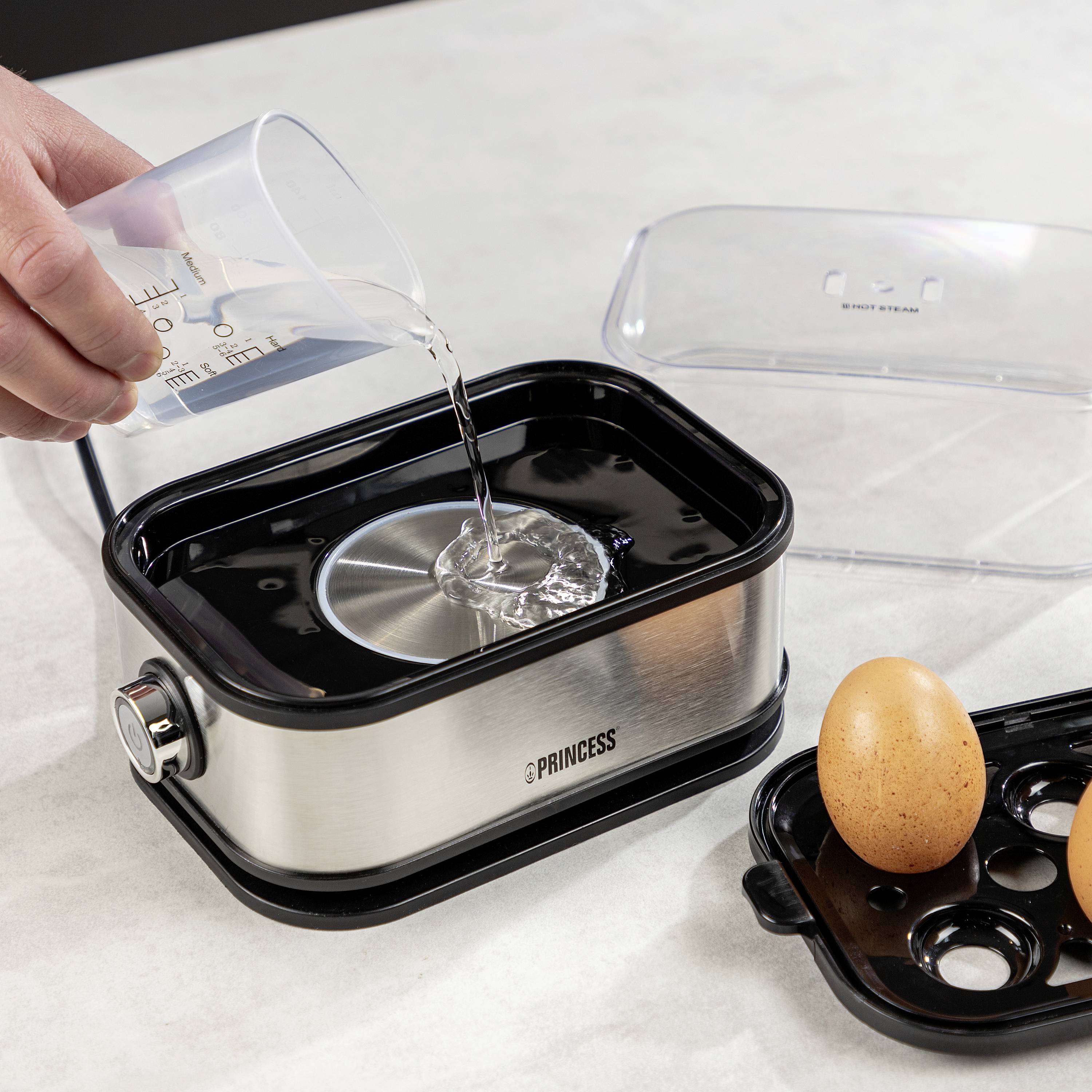 Een roestvrijstalen eierkoker, waarin water wordt gegoten uit een maatbeker; ernaast twee eieren in zwarte houders.