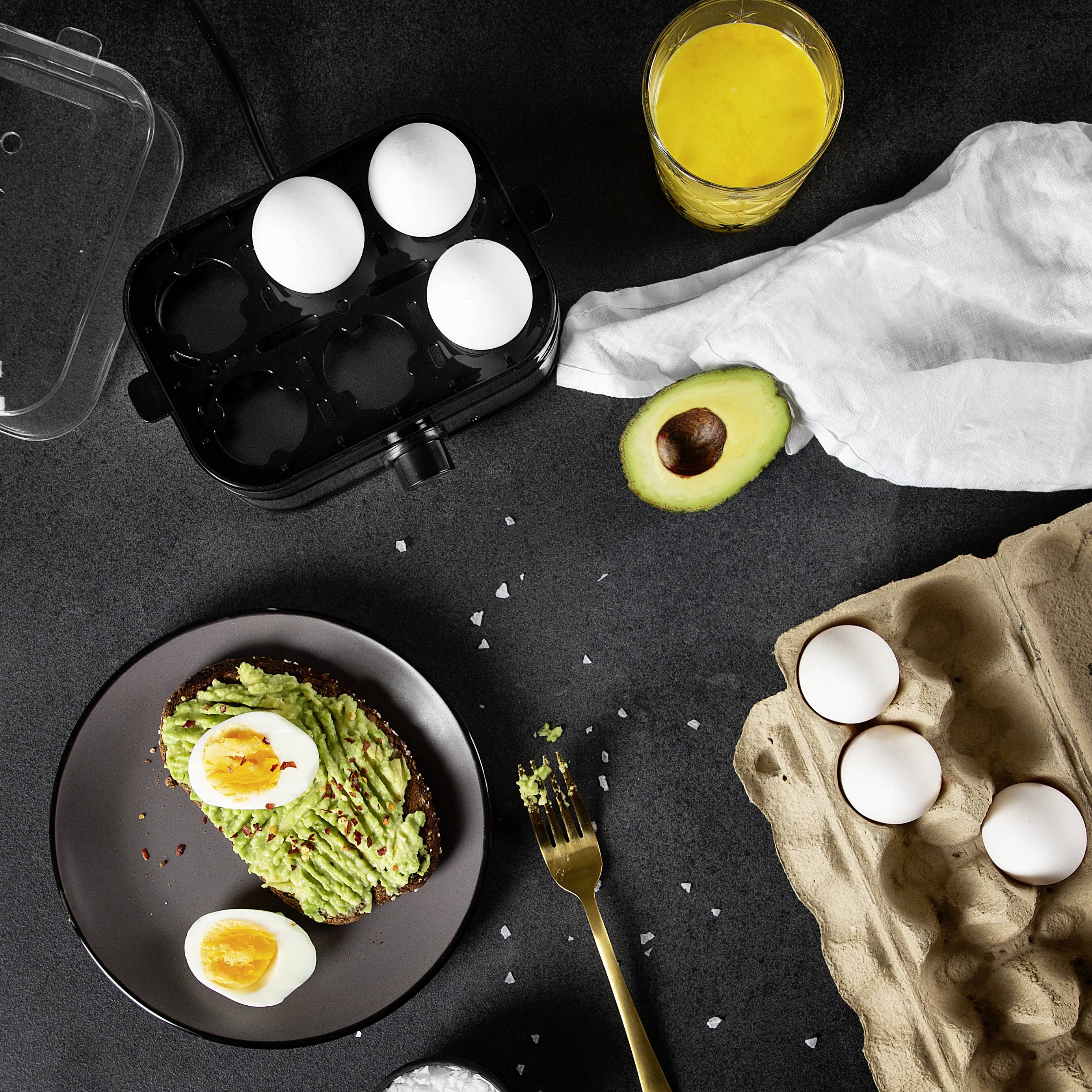 Avocado-toast met gekookt ei op een zwarte schaal, naast gesneden avocado, rauw ei, eierdoos en een glas sinaasappelsap op een donkere tafel.