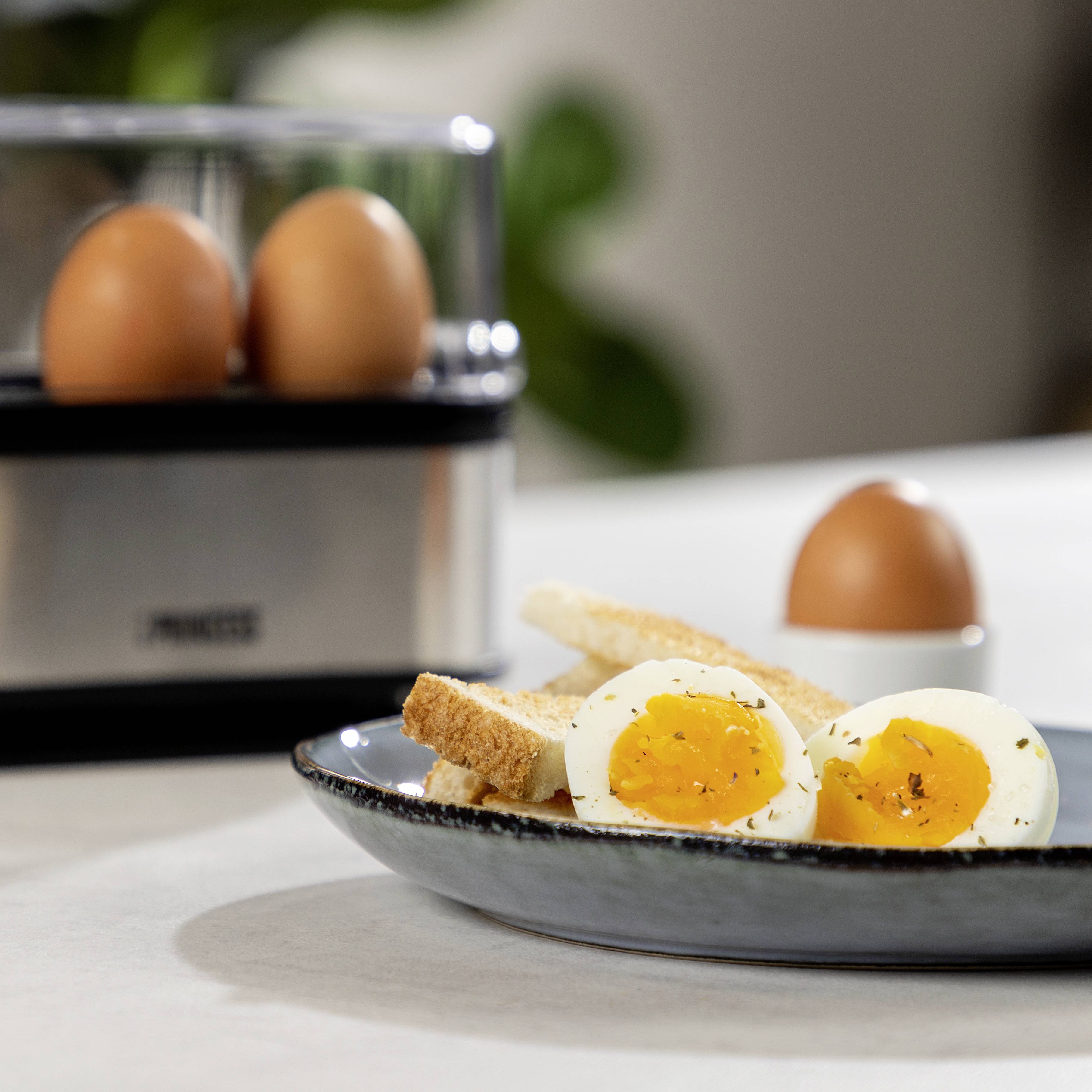 Een bord met gesneden, gekookte eieren en toaststrips. Op de achtergrond een eierkoker met twee eieren.