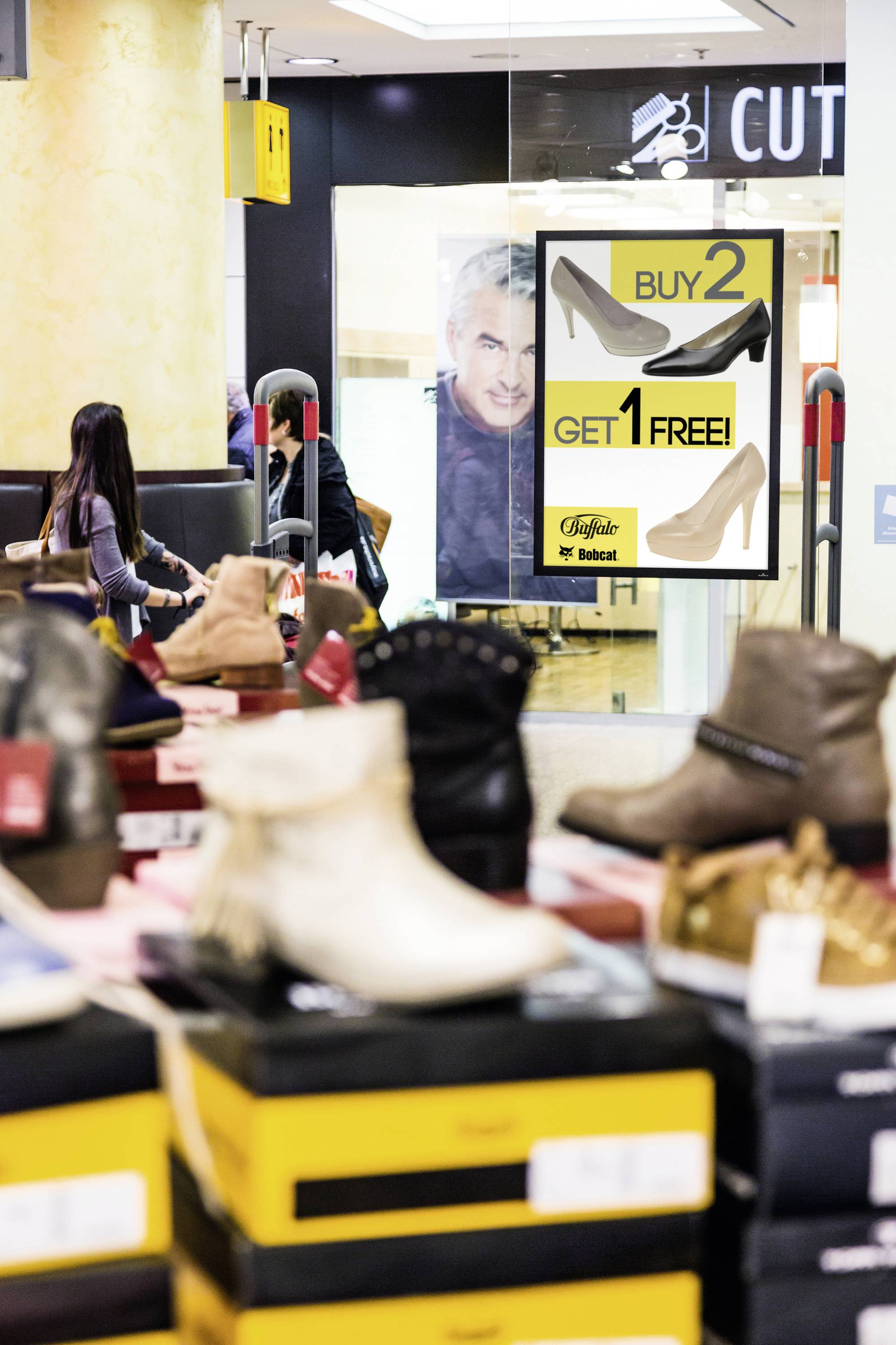 Een schoenwinkel met een reclameposter op de achtergrond: 'Koop 2, Krijg 1 Gratis'. Verschillende schoenen op tafels op de voorgrond.