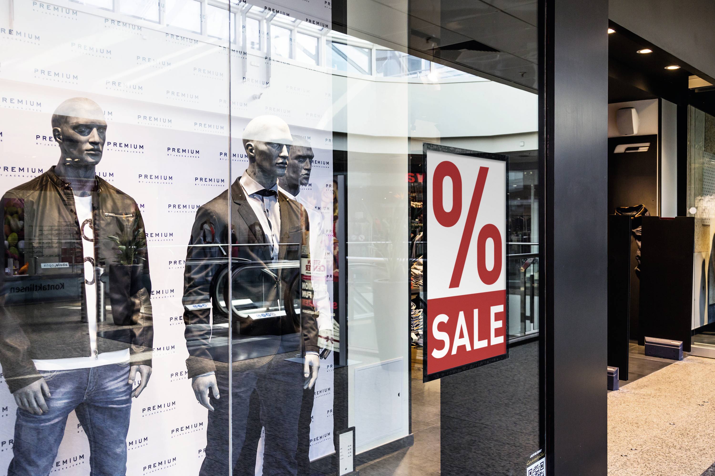 Etalage van een kledingwinkel met twee etalagepoppen in zwarte jassen. Een groot bord toont '% SALE'.