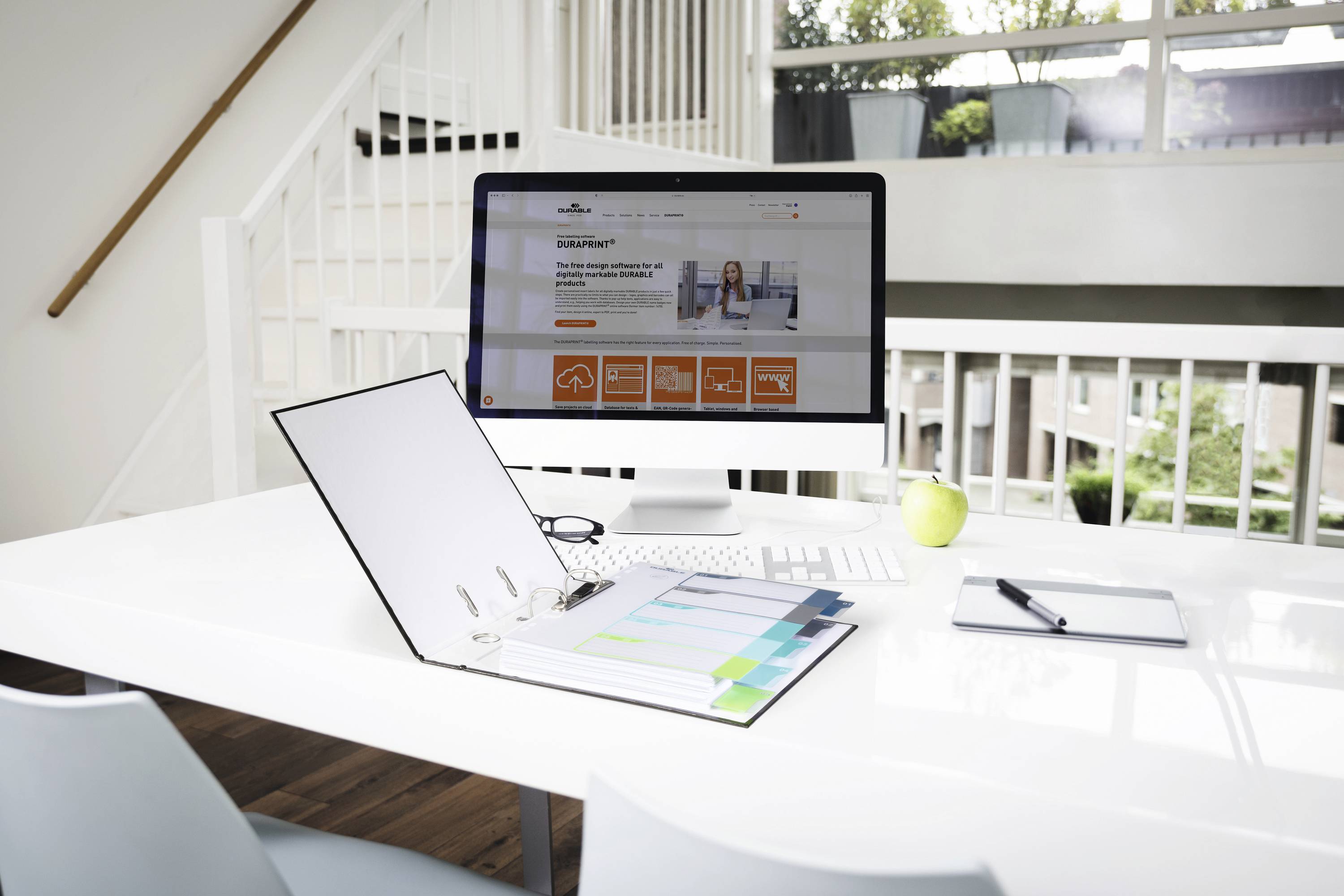 Een opgeruimd bureau met computer, opengeslagen map met tabbladen, notitieboekje, pen en appel. Grote ramen op de achtergrond.