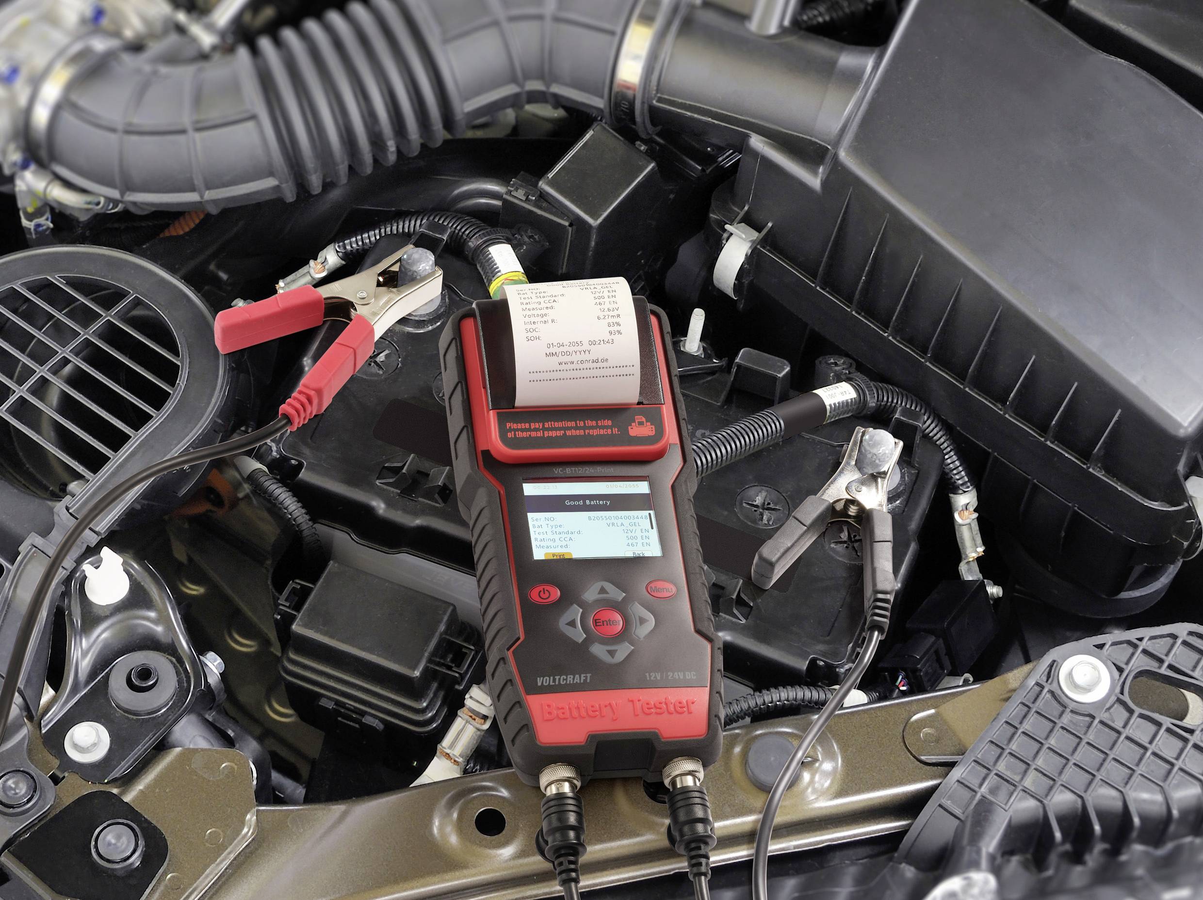 Een batterijtest wordt uitgevoerd met een batterijmeter die is aangesloten op een autoaccu in de motorruimte en meetgegevens weergeeft op het display.