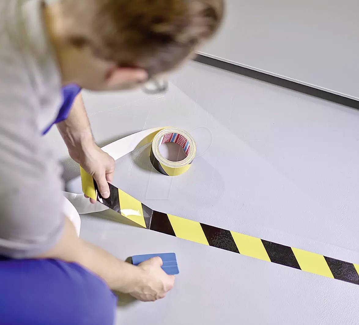 Een persoon plakt een geel-zwart waarschuwingslint op een grijze vloer. Op de achtergrond ligt een rol tape.