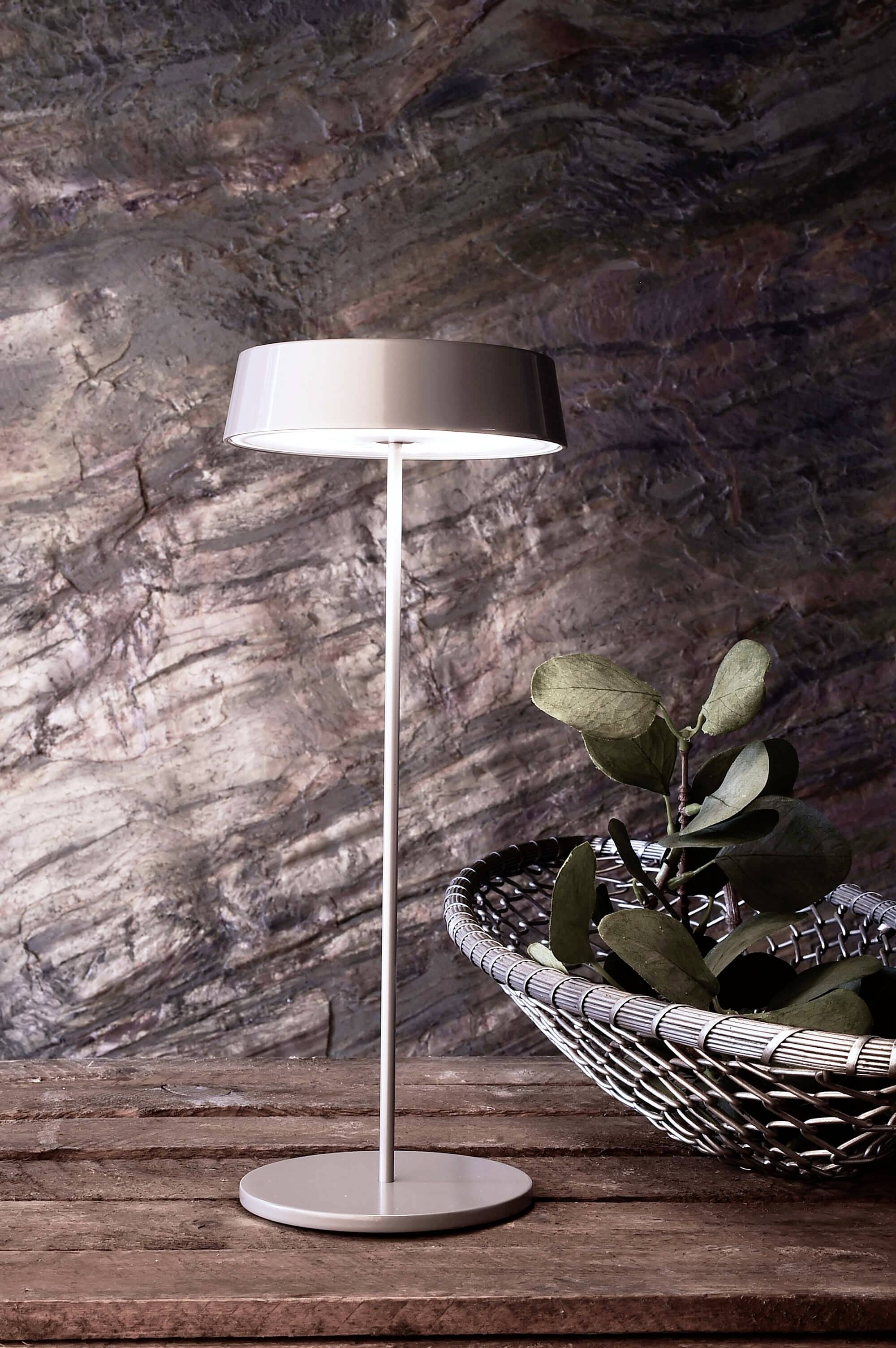 Staande lamp met modern design naast een gevlochten schaal met groene bladeren op een houten tafel voor een stenen muur.