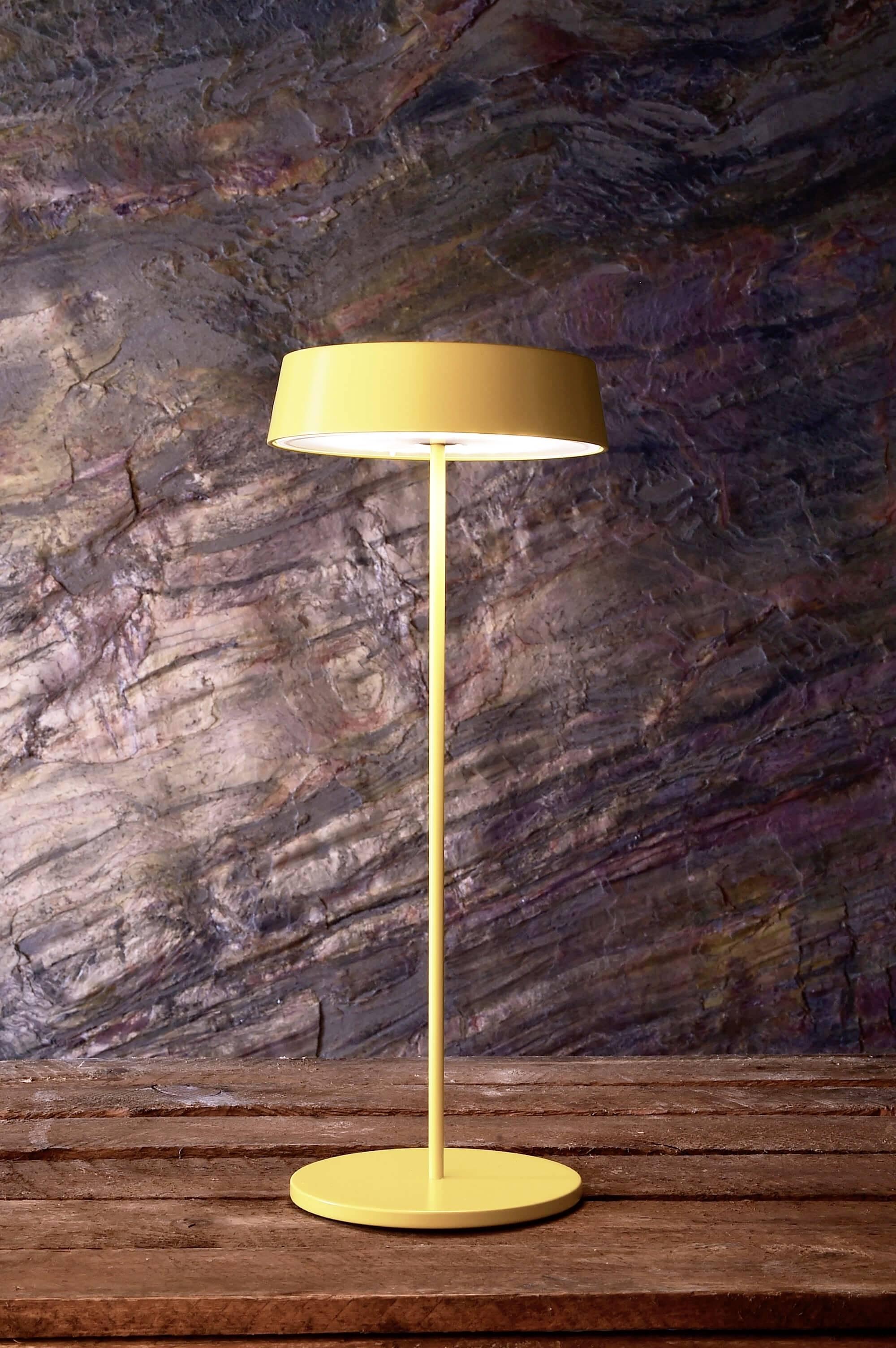 Gele staande lamp met een ronde kap voor een gestructureerde muur op een houten tafel. De lamp verspreidt zacht licht.