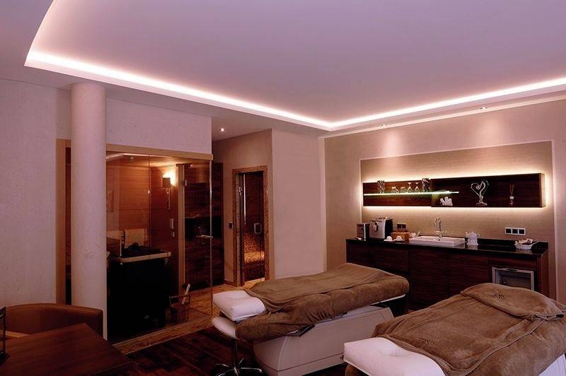 Behandelruimte in een spa met twee massagetafels, zachte verlichting en een sauna op de achtergrond. Een rustige en ontspannende sfeer.