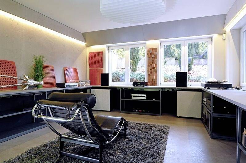 Een modern woonkamer met een zwarte leren fauteuil op een vloerkleed. Grote ramen laten daglicht binnenstromen, stereoapparatuur staat opgesteld op een rek links.