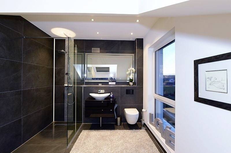 Modern badkamer met zwarte tegelmuur, inloopdouche en wastafel, grote spiegel, witte toiletpot en raam.