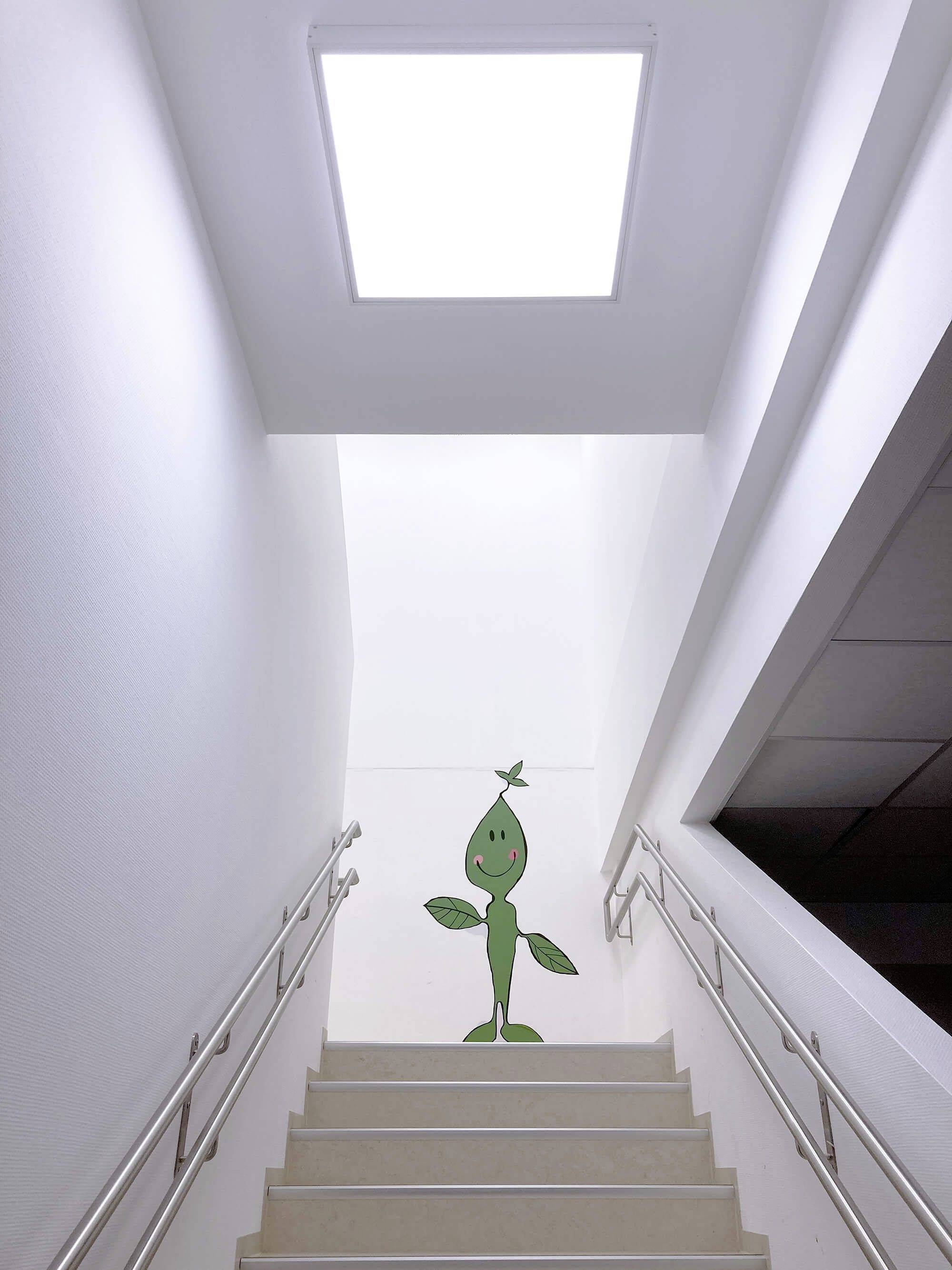 Een groene, cartoonachtige plant met een gezicht staat bovenaan een trap in een modern gebouw, daaronder een wit verlicht plafondpaneel.