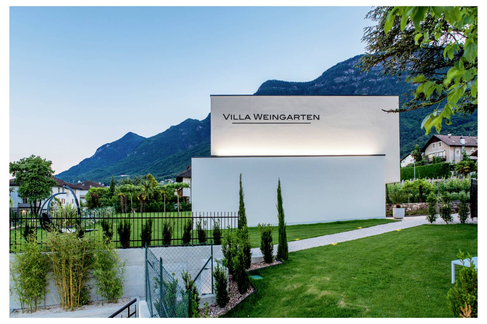Modern wit gebouw met het opschrift 'VILLA WEINGARTEN' naast een verzorgd gazon, met bergen en huizen op de achtergrond bij zonsondergang.